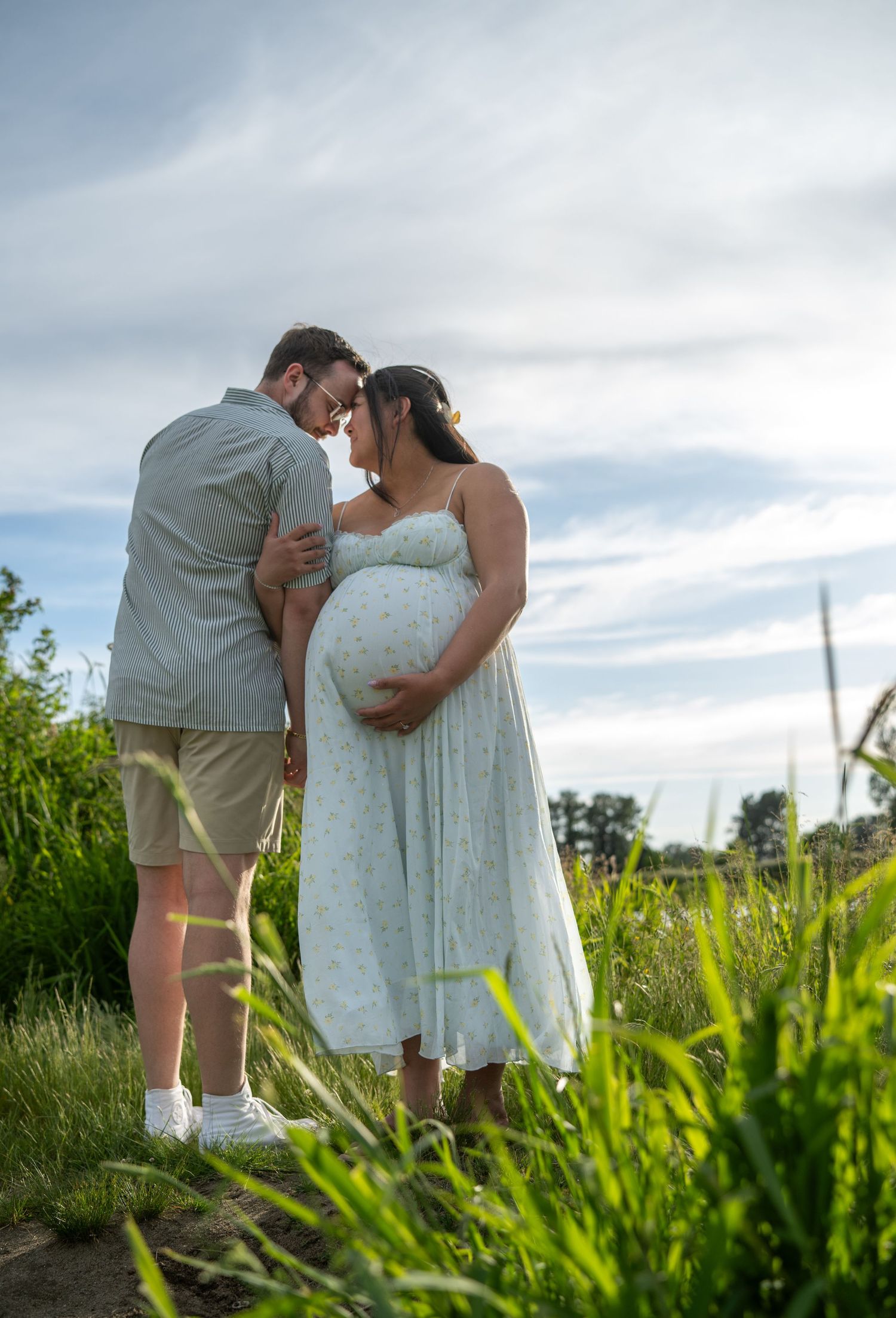 Expectant couple sharing a tender forehead touch in a lush green field, woman in floral maternity dress.