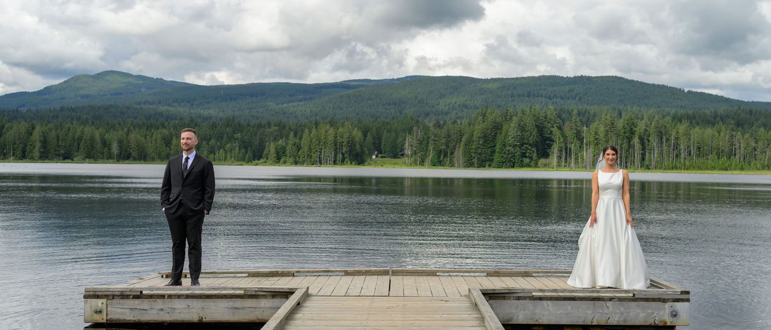 Bride and groom stand apart on a wooden dock overlooking a serene mountain lake surrounded by lush forest.