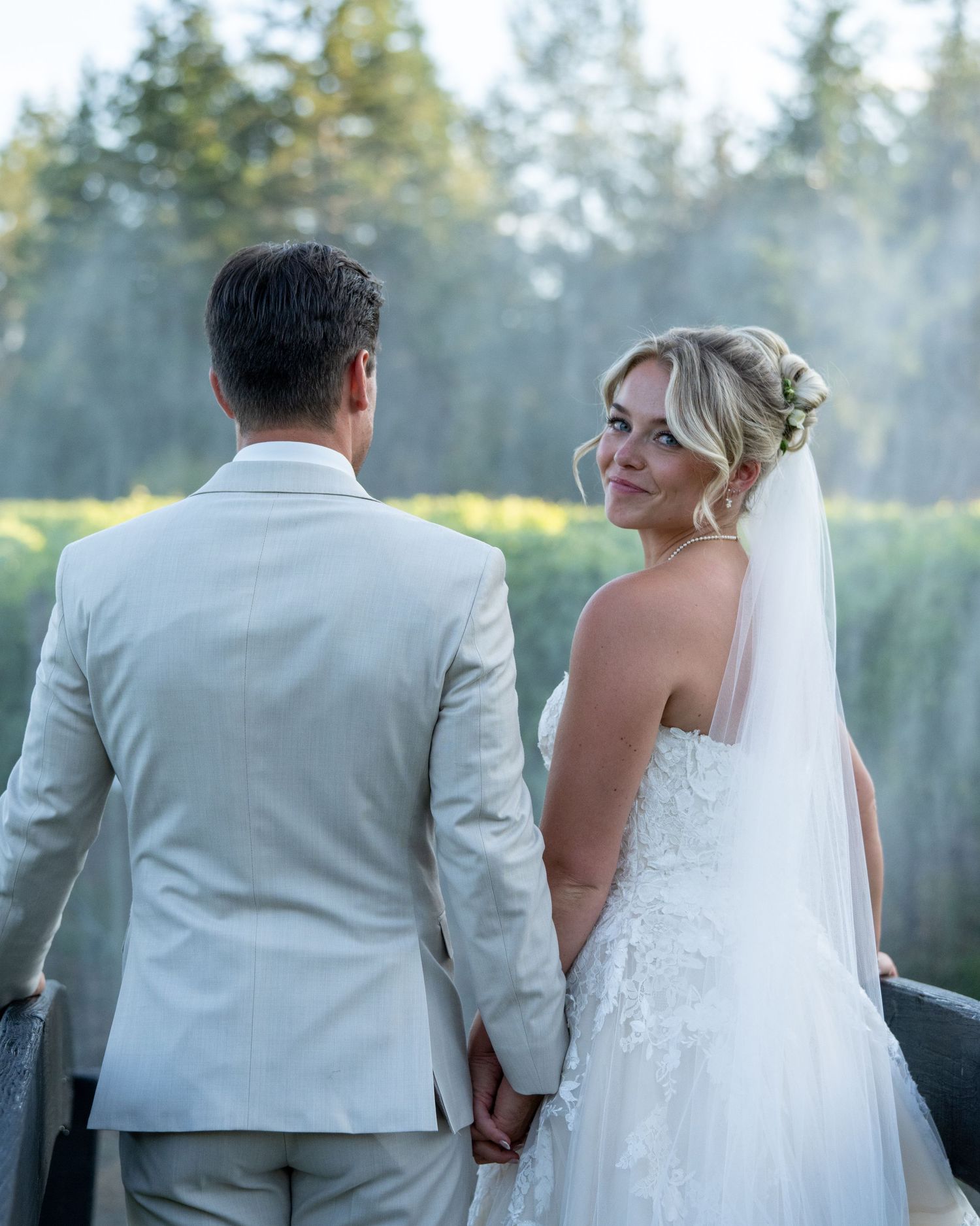 Bride in lace gown and veil holds hands with groom in white suit, glancing back during outdoor wedding.