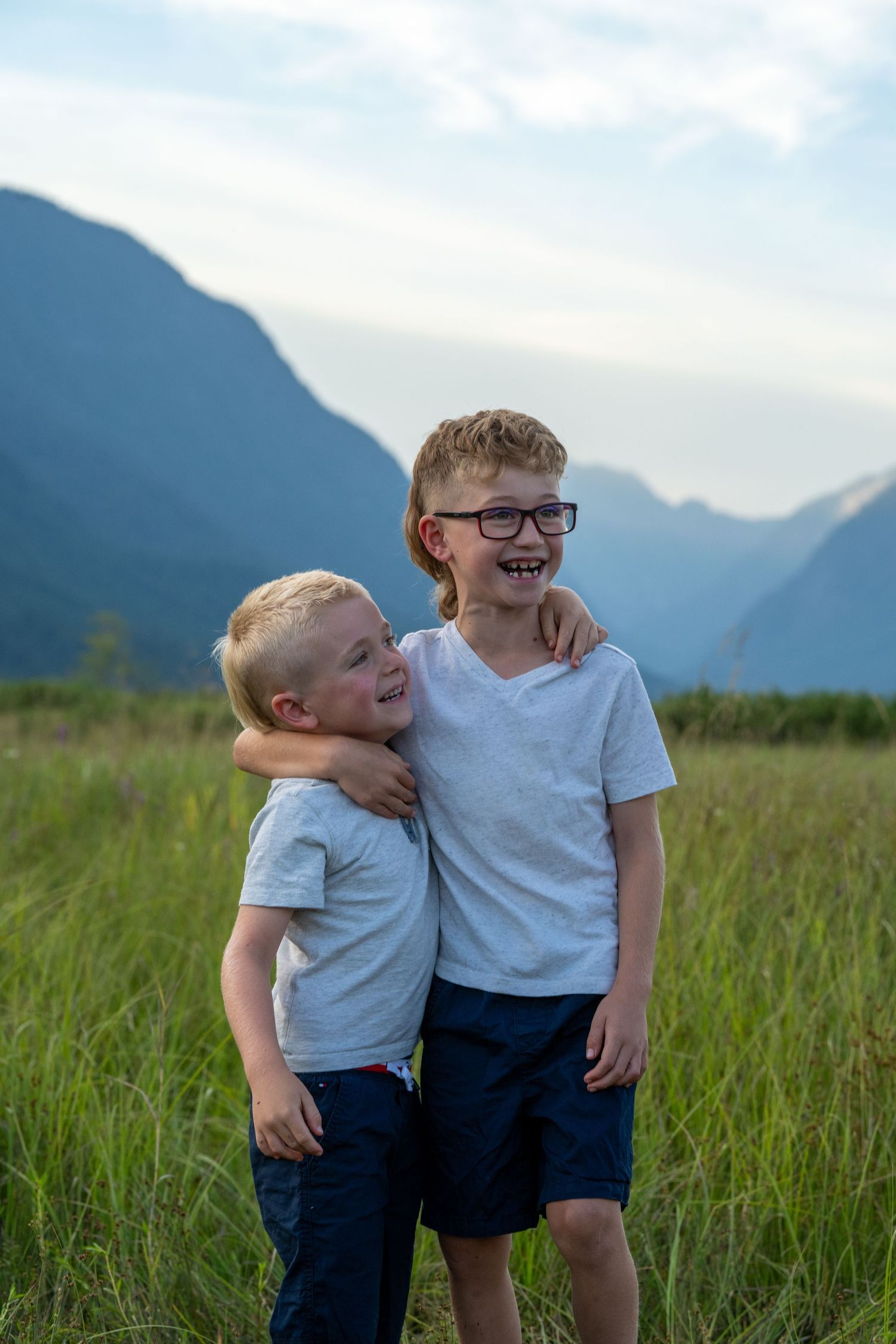 Two smiling boys in white t-shirts embrace in a green meadow with mountain backdrop.