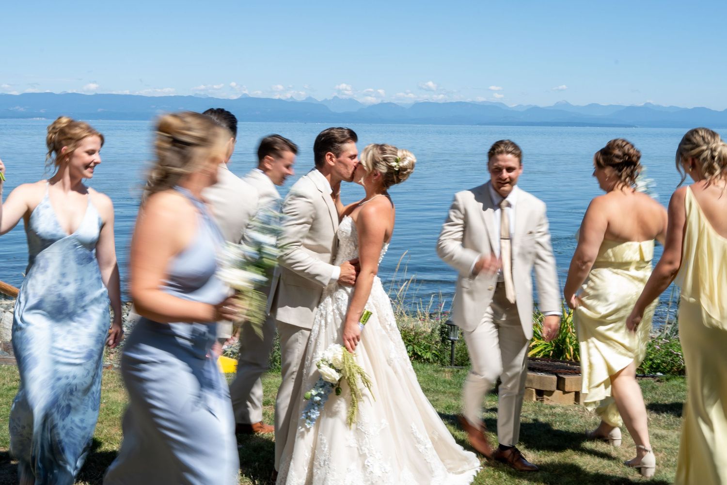 Bride and groom share a kiss surrounded by wedding party in blue and yellow dresses by a scenic lakeside.