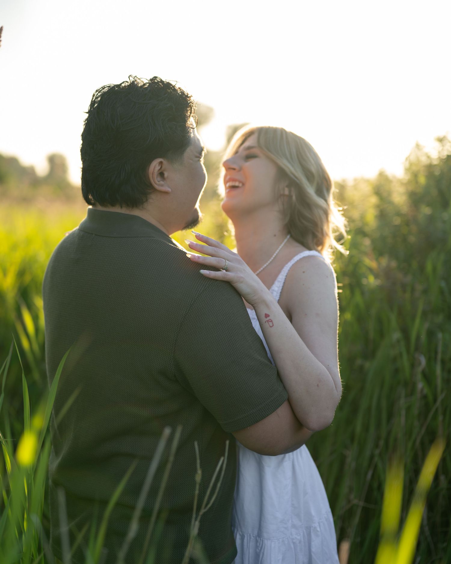 Couple embracing romantically in a lush green field at golden hour, woman in white dress smiling up at man in olive shirt.