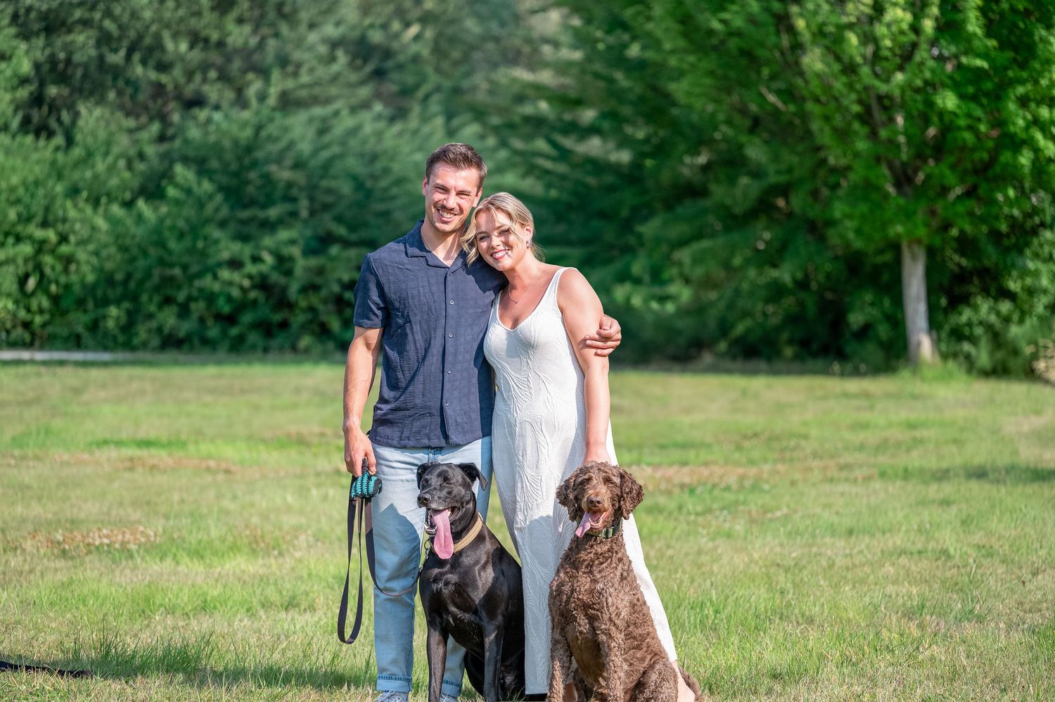A couple stands outdoors in a grassy field with their two dogs, a grey one and a brown one, on a sunny day. Colony Farms Park Port Coquitlam
