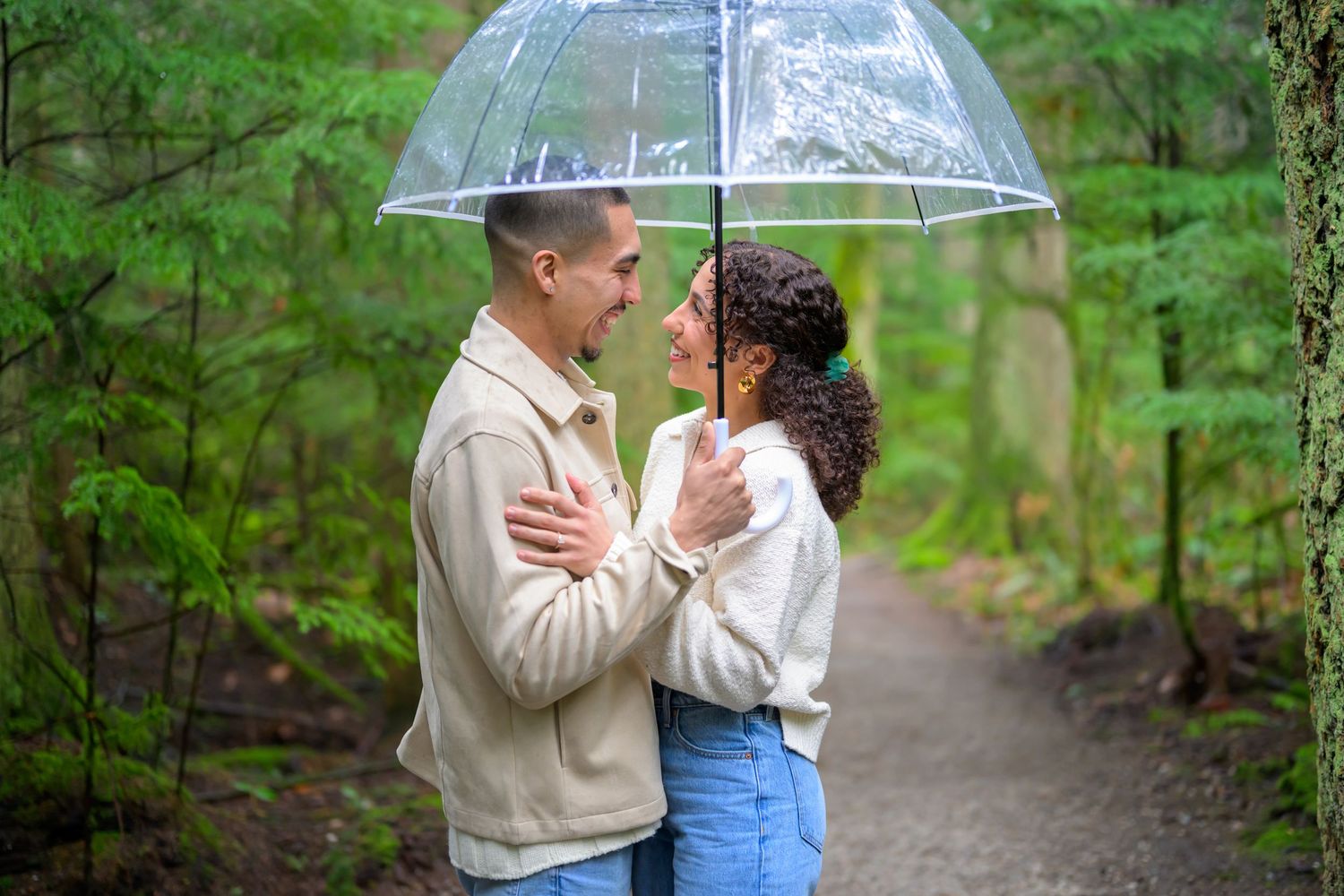Couple sharing tender moment under clear umbrella on rainy forest path.
