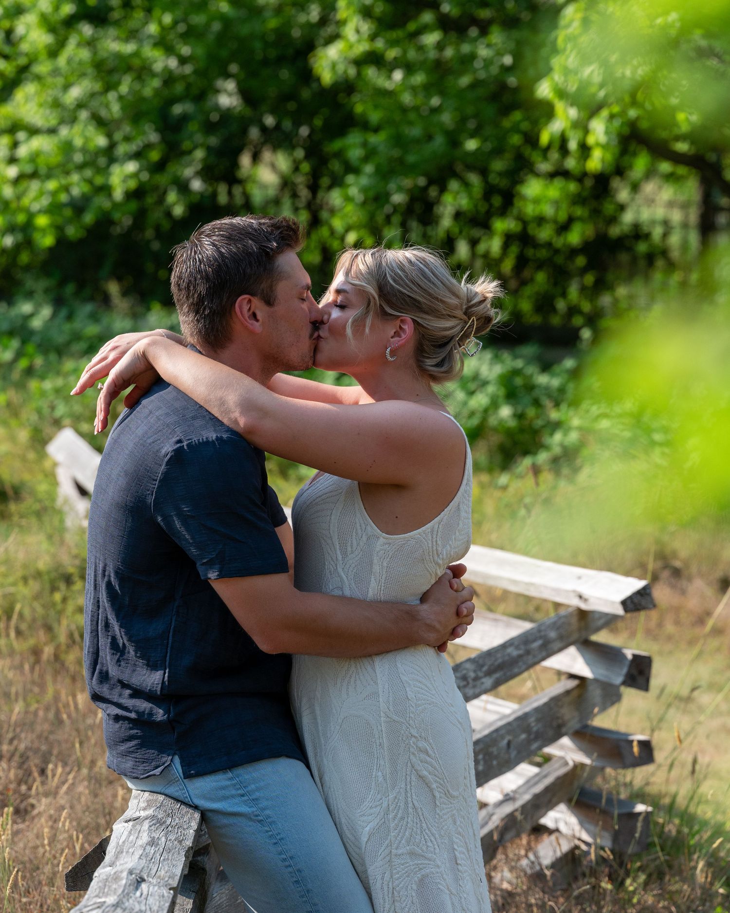 A couple embraces near a rustic wooden bench surrounded by lush greenery.