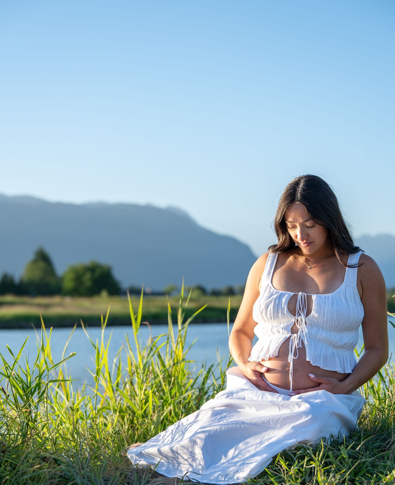 Pregnant woman in white dress sitting on grass by a lake, cradling her baby bump outdoors.