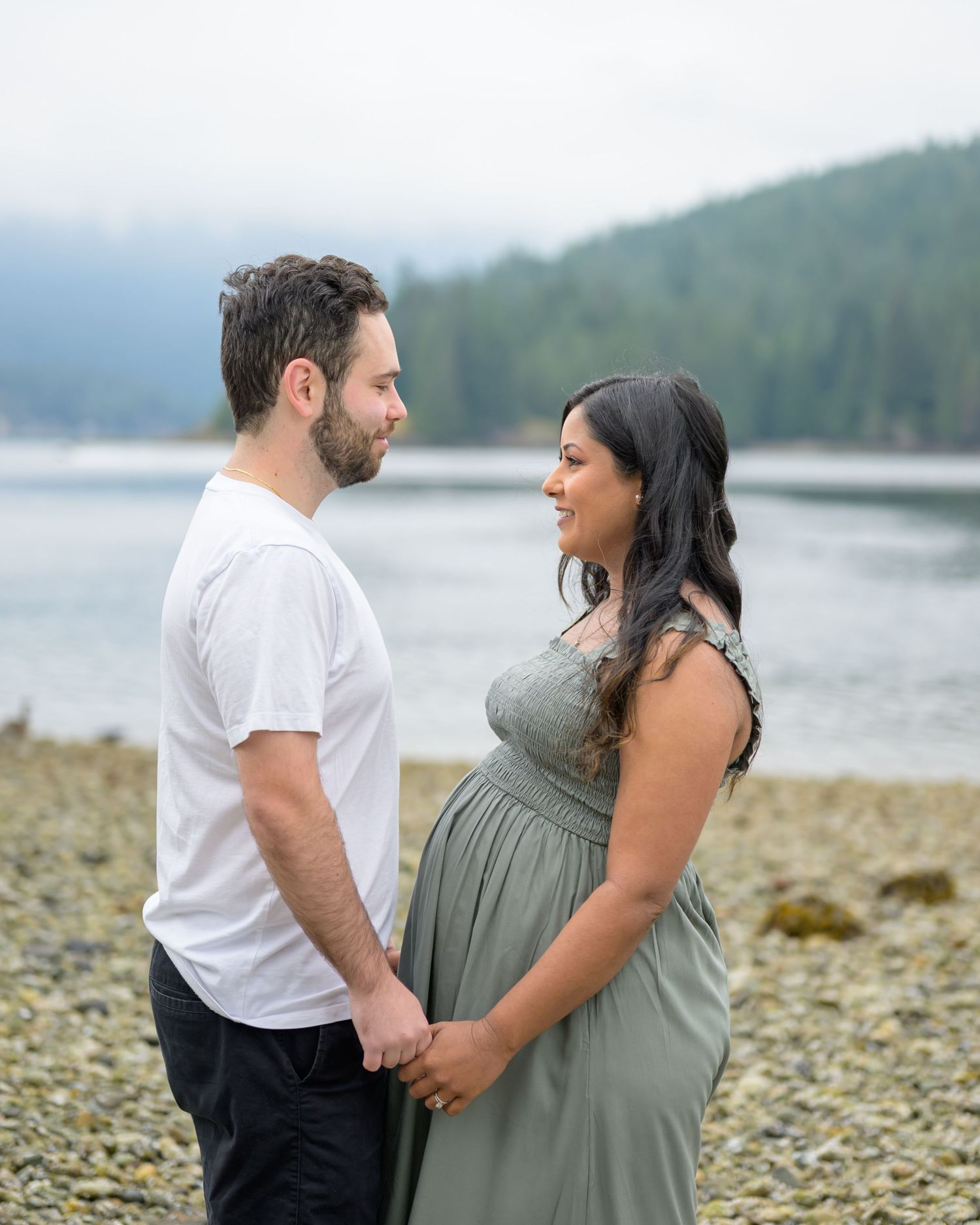 Pregnant couple holding hands on rocky beach with misty mountains and calm water in background.