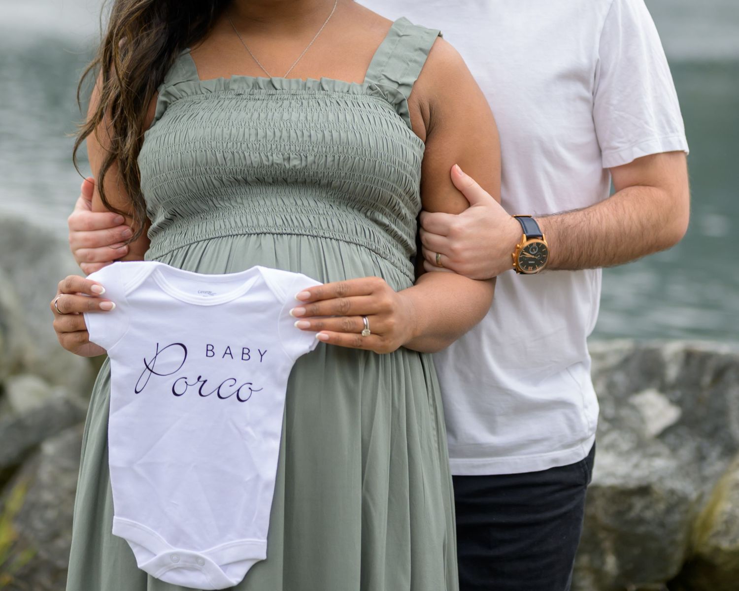 Pregnant couple holding a white Baby Porco onesie outdoors near water, woman in sage green dress.