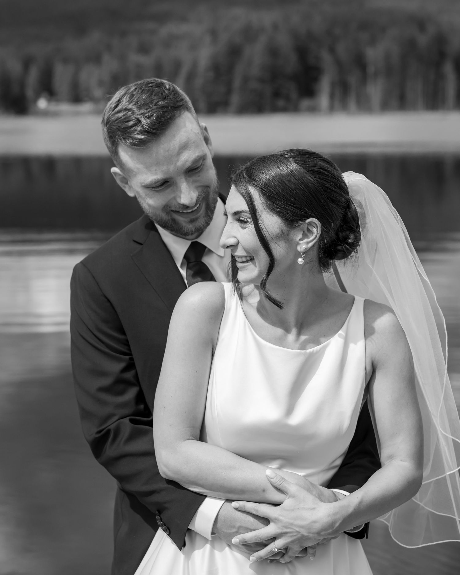 Black and white photo of a smiling bride and groom embracing near a lake, bride wearing veil and white dress.