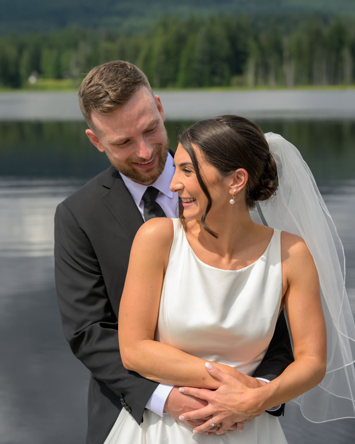 Bride and groom embrace lakeside, she in white gown with veil, he in dark suit, with forested mountains behind.