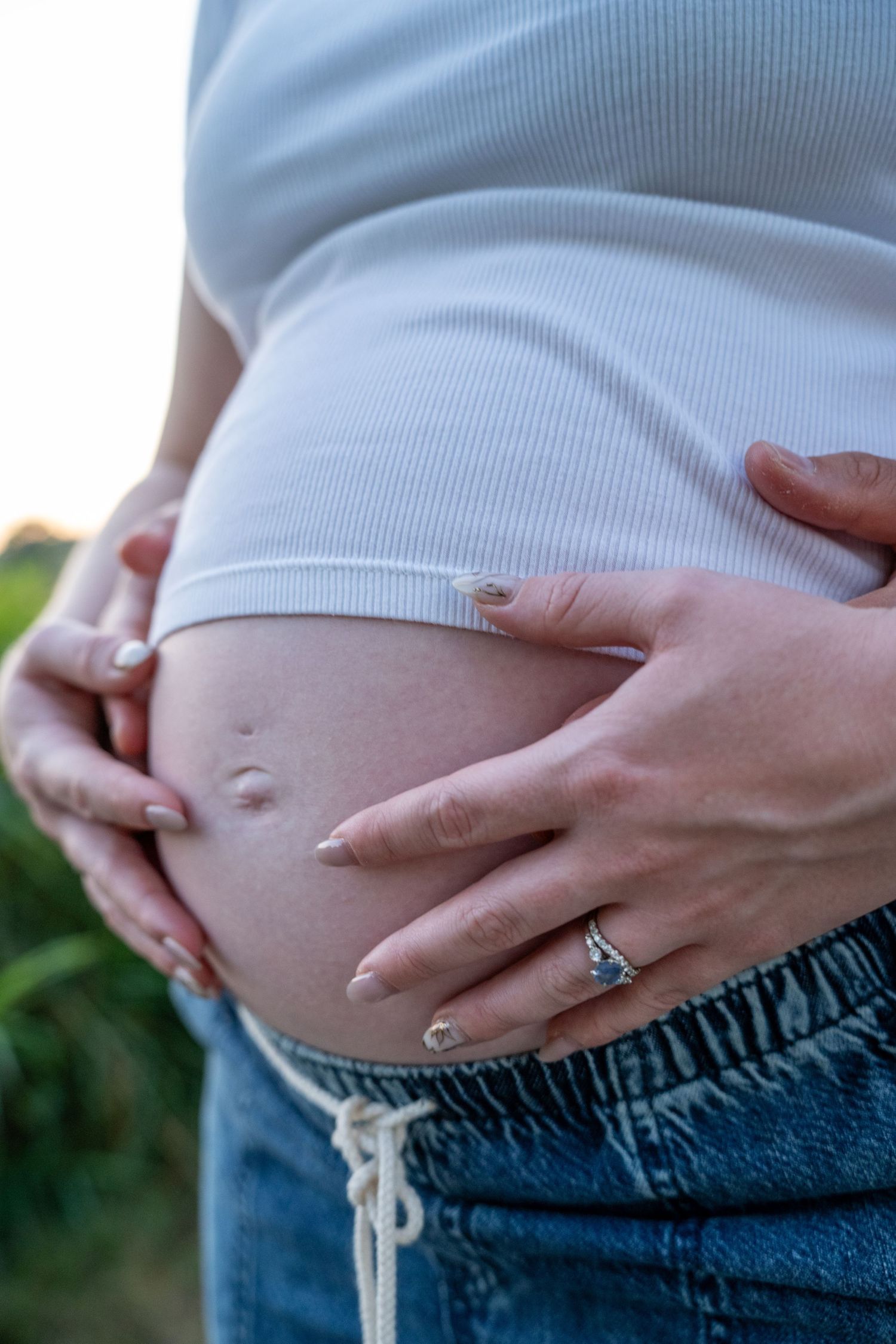 Pregnant woman in white crop top and jeans cradling her baby bump outdoors, wearing a ring on her finger.