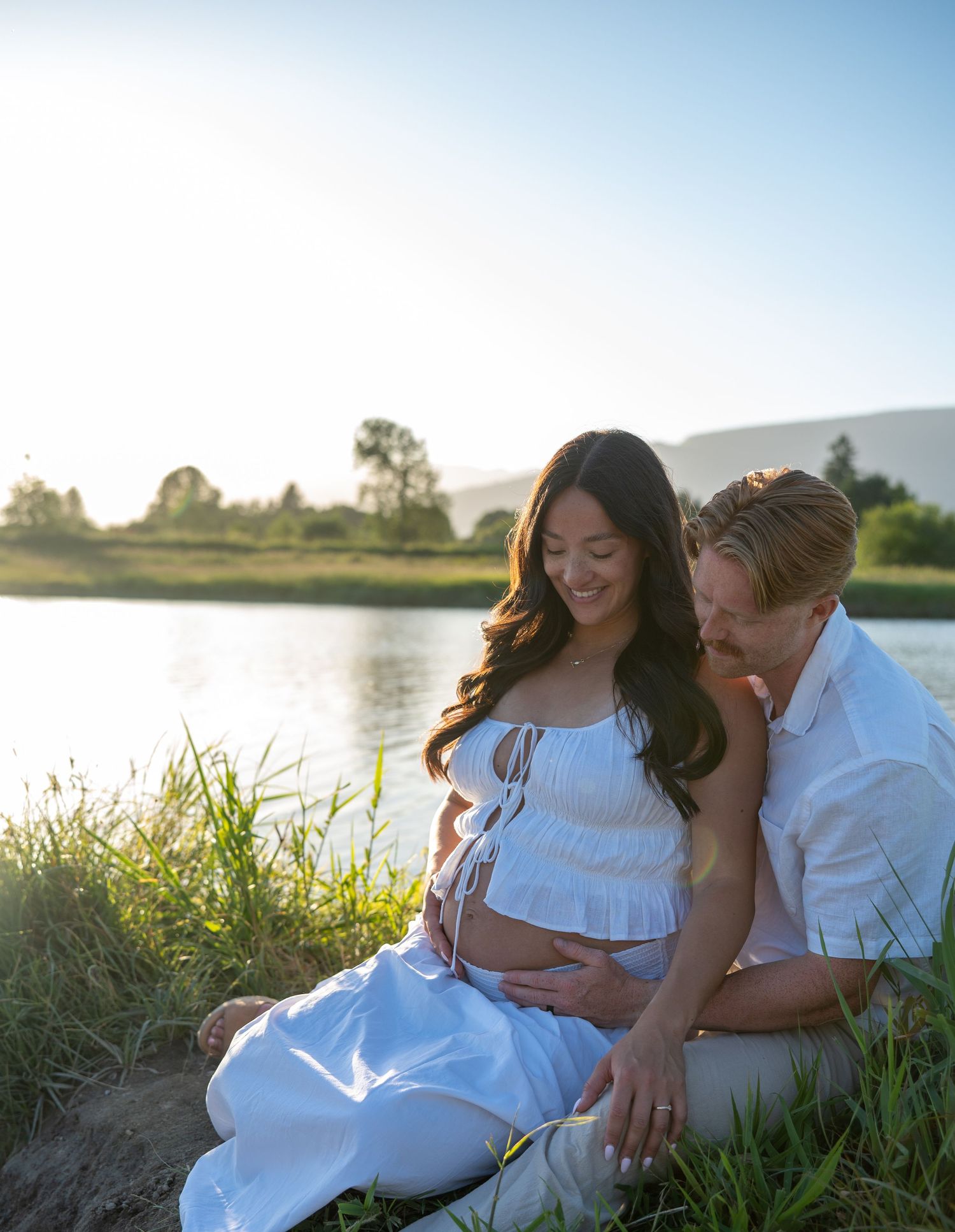 Pregnant couple sitting by a sunlit river, woman in white dress cradling baby bump, man tenderly leaning in beside her.