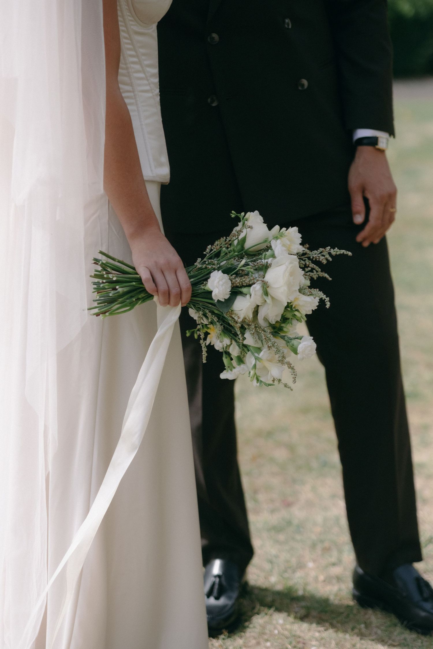 Bride holding white floral bouquet in elegant wedding gown beside groom in black suit outdoors.