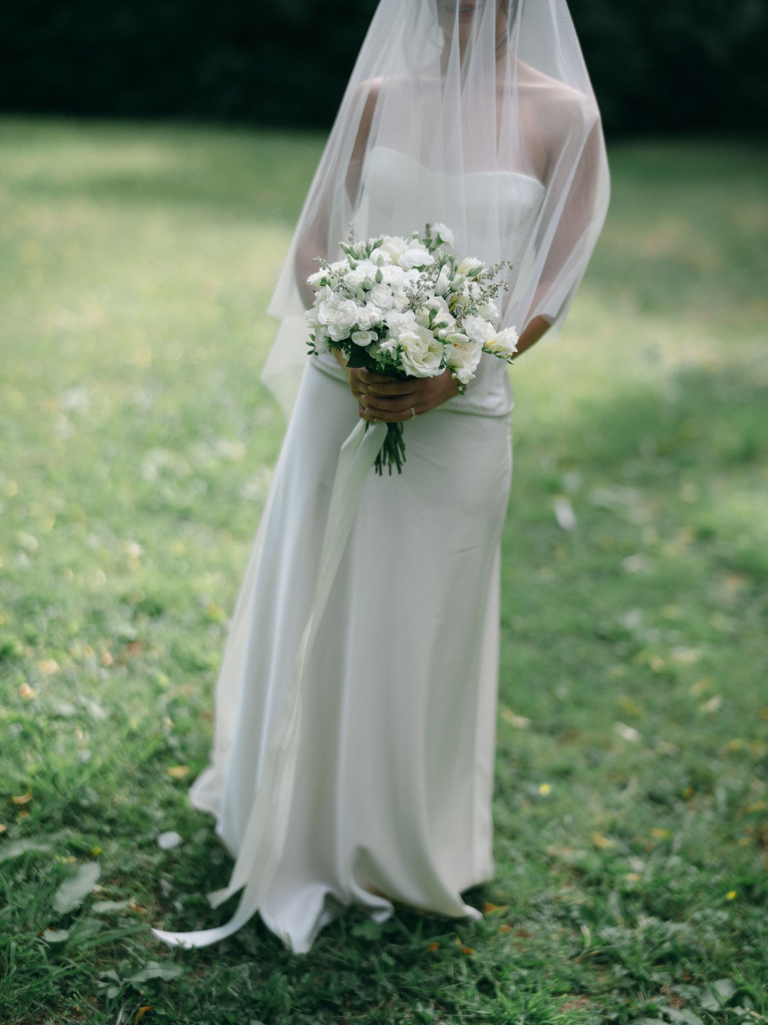 Bride in a strapless white gown and veil holds a white rose bouquet on a green lawn.