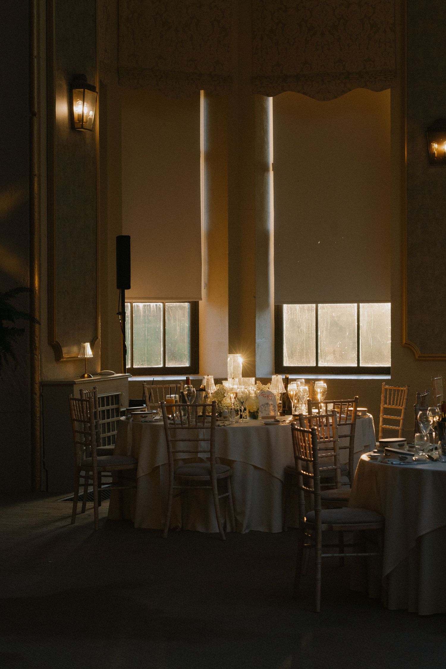 Elegant dimly lit banquet hall with round tables, chiavari chairs, candles, and tall windows for a wedding reception.