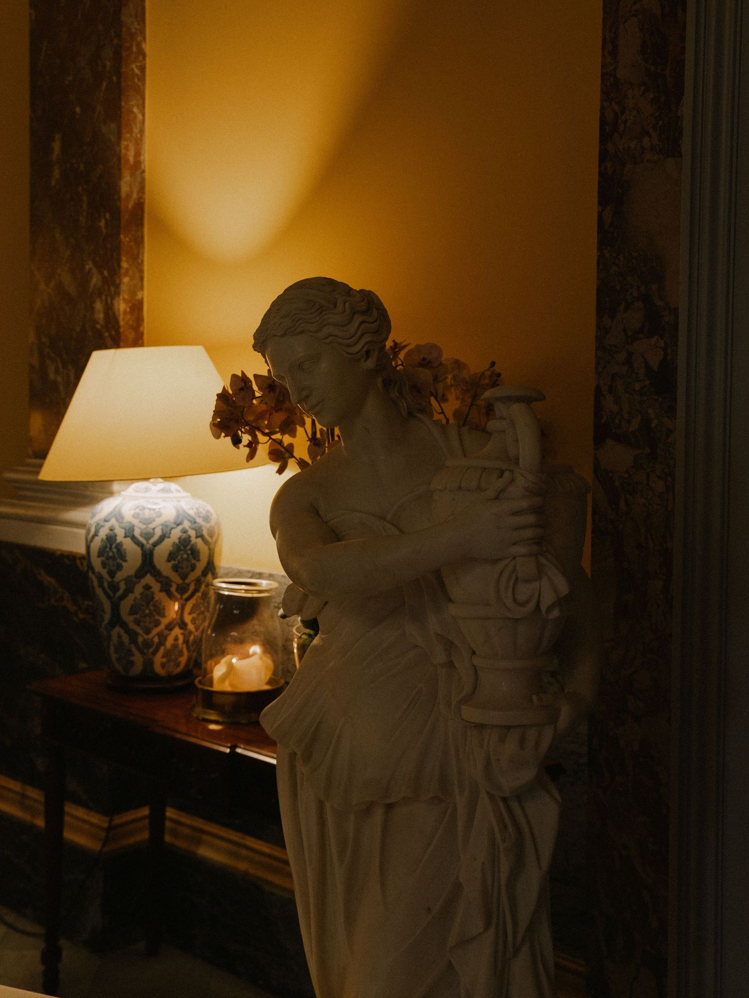 Classical white marble female statue beside a glowing table lamp with blue ceramic base in warm golden interior.