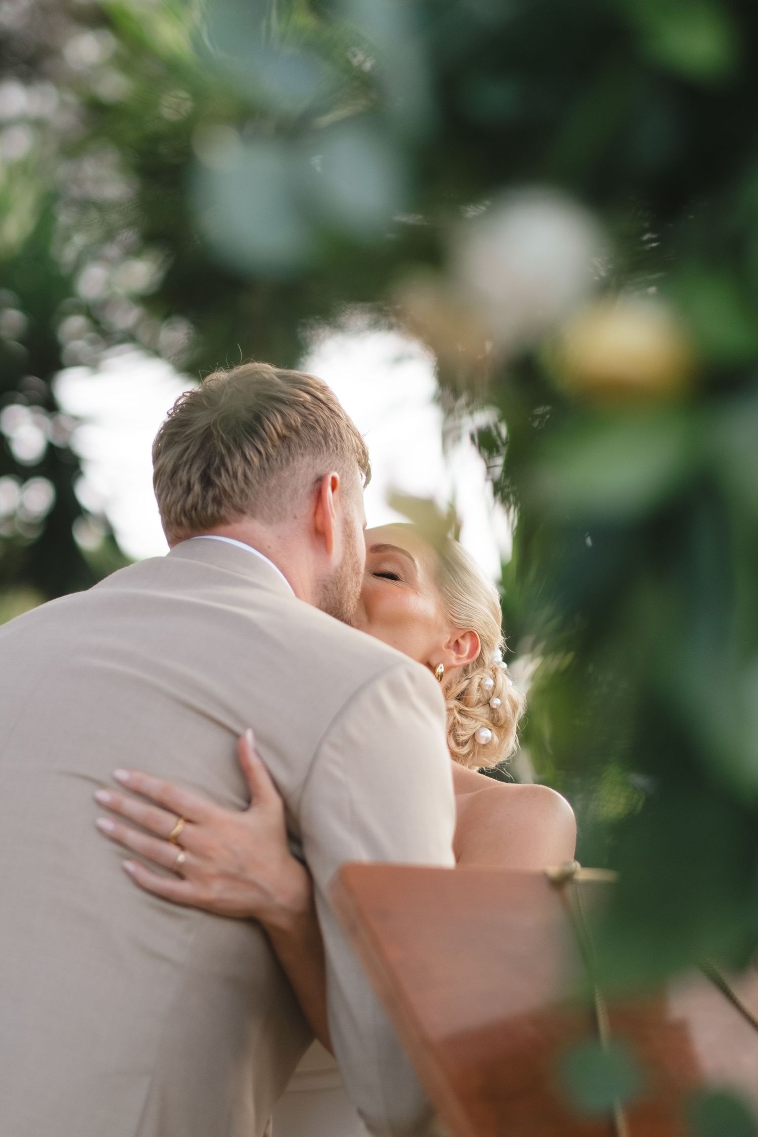 Bride and groom sharing tender first kiss embrace surrounded by lush greenery.