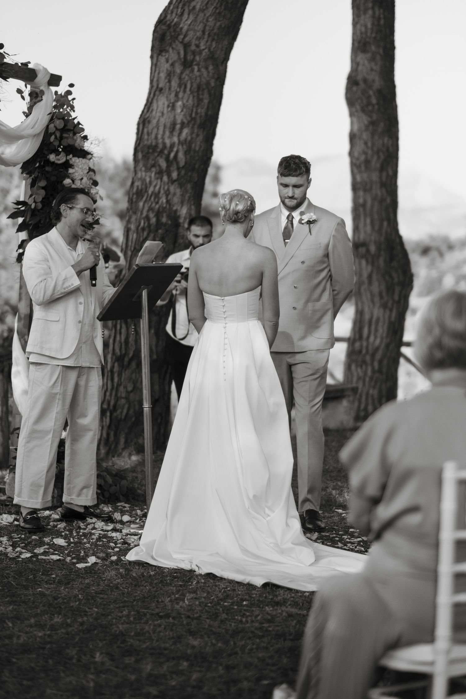 Black and white photo of bride and groom exchanging vows at outdoor wedding ceremony with officiant.