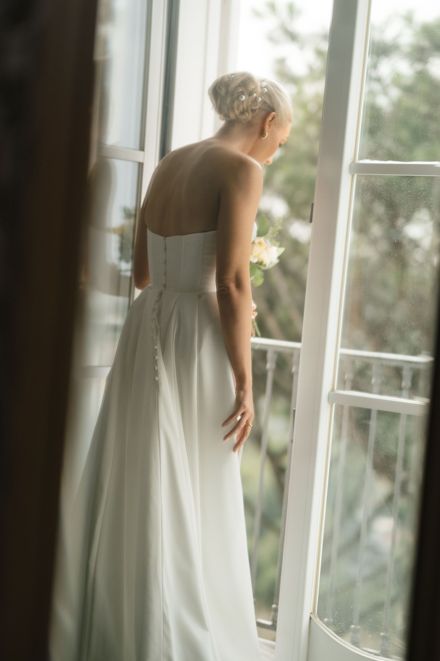 Bride in strapless white gown holding bouquet, gazing out French doors with soft natural light.