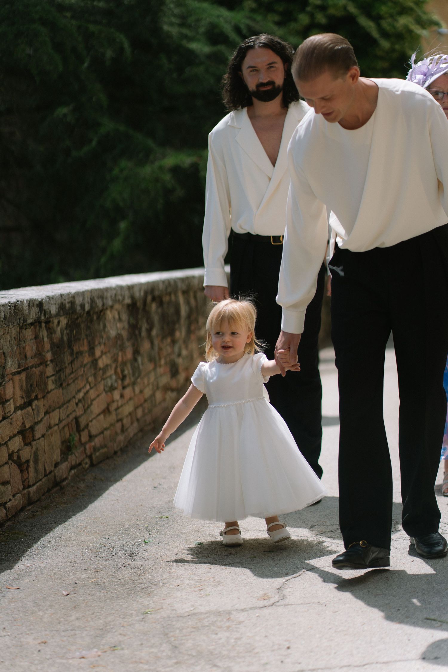 Adorable blonde flower girl in white dress holding hands with a man in white shirt at an outdoor wedding ceremony.