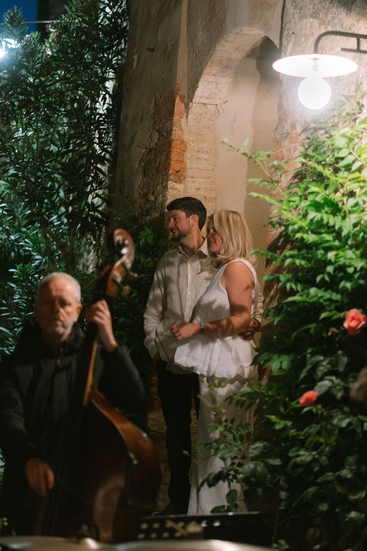 Couple sharing a romantic moment in a candlelit garden courtyard while a musician plays double bass nearby.