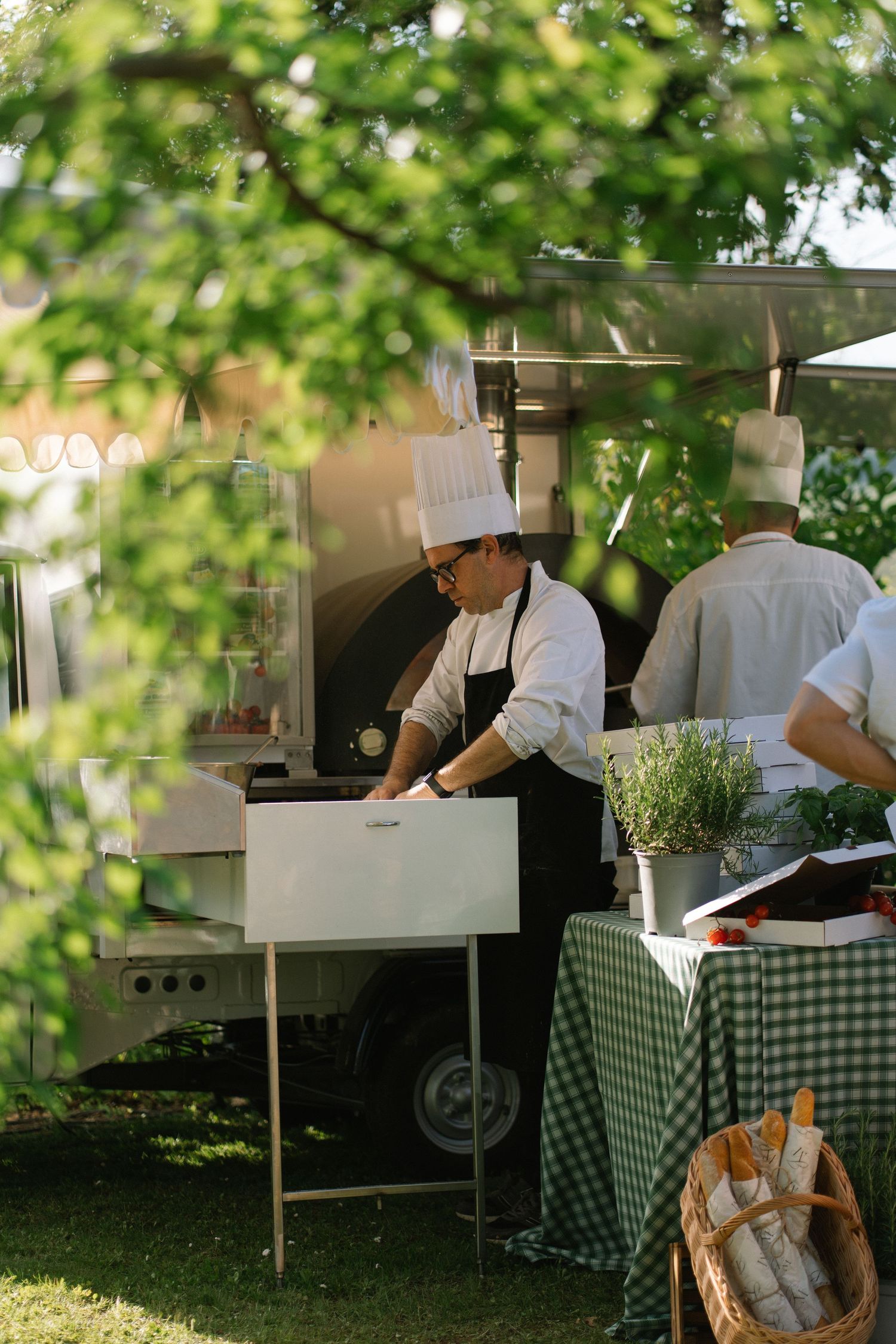 Chef in white hat working at an outdoor kitchen station beside a mobile pizza oven.