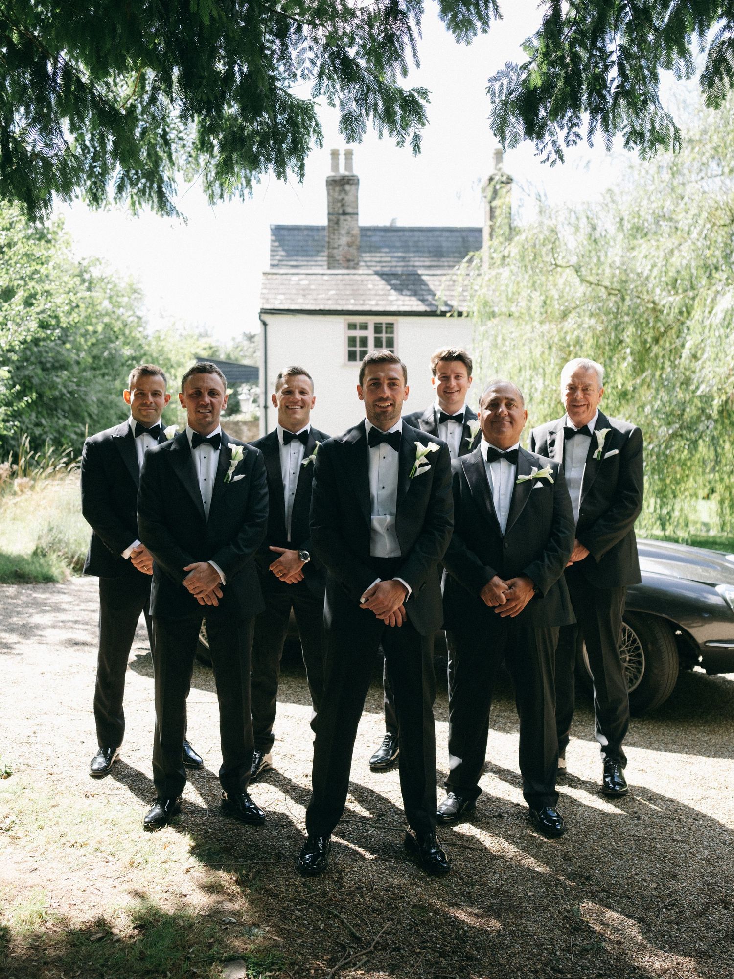 Groom and groomsmen in black tuxedos with bow ties posing outdoors near a country house on a sunny wedding day.