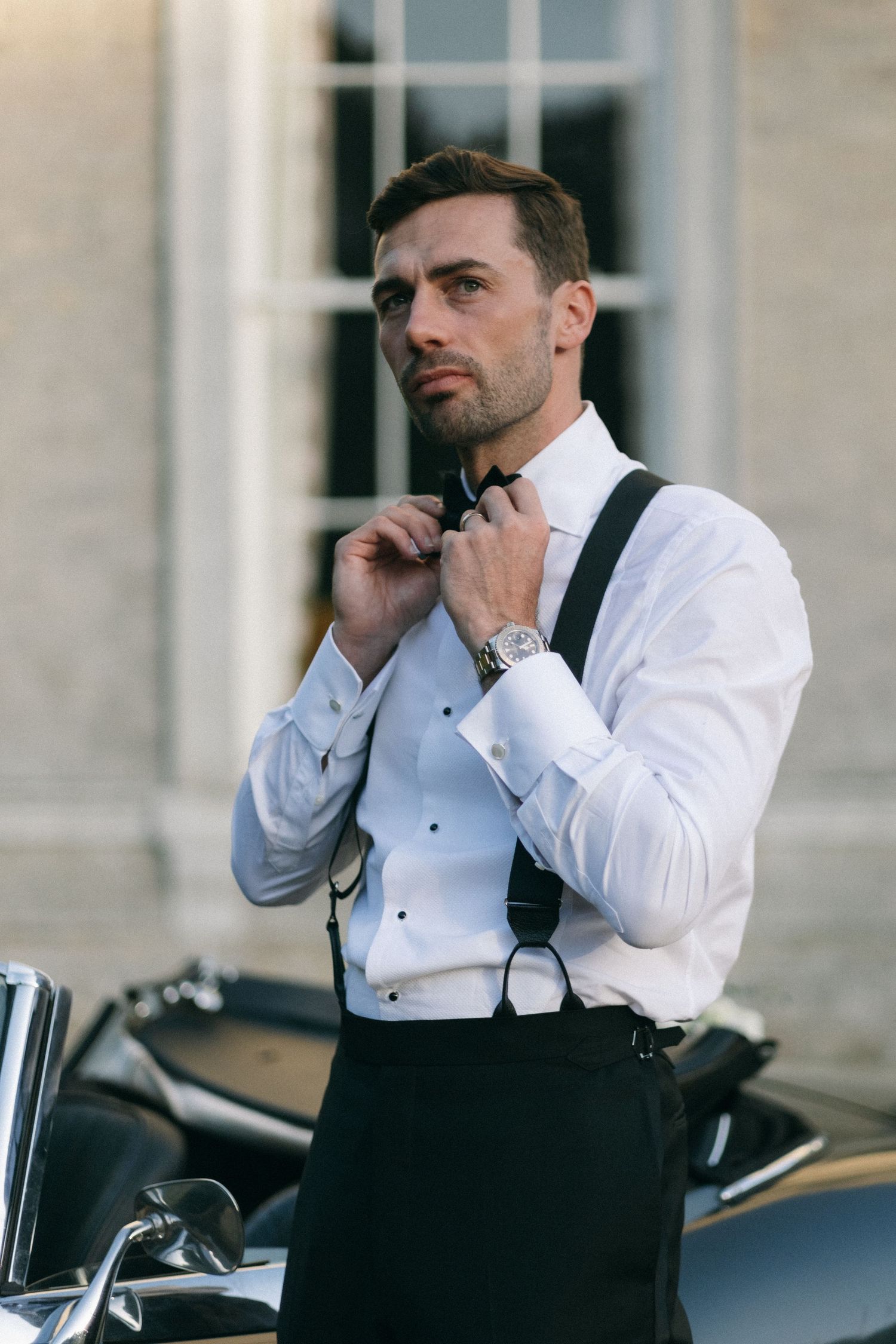 Elegant man in white tuxedo shirt adjusting bow tie with suspenders, standing beside classic car.