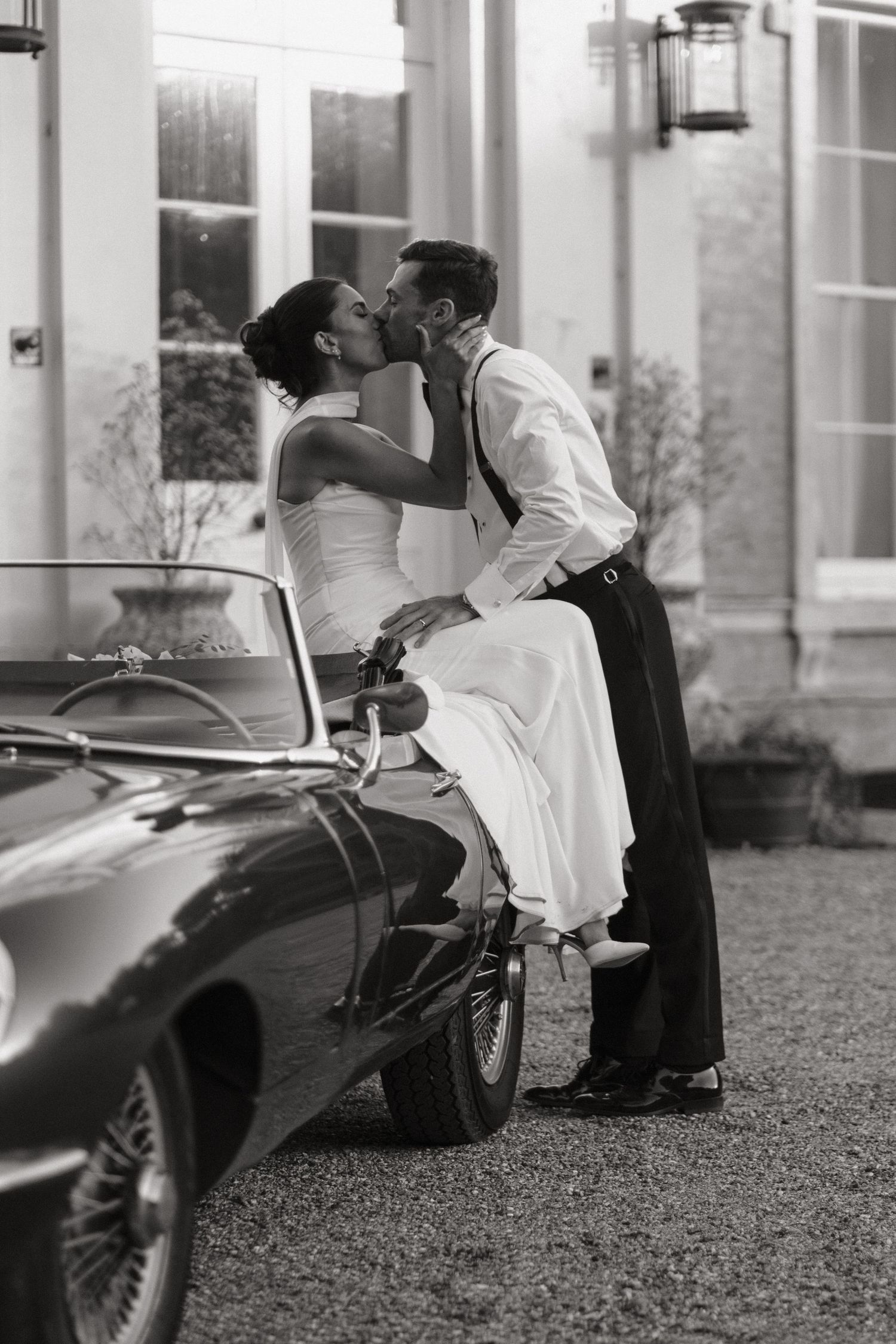 Black and white photo of a bride and groom kissing beside a vintage convertible car outside an elegant venue.
