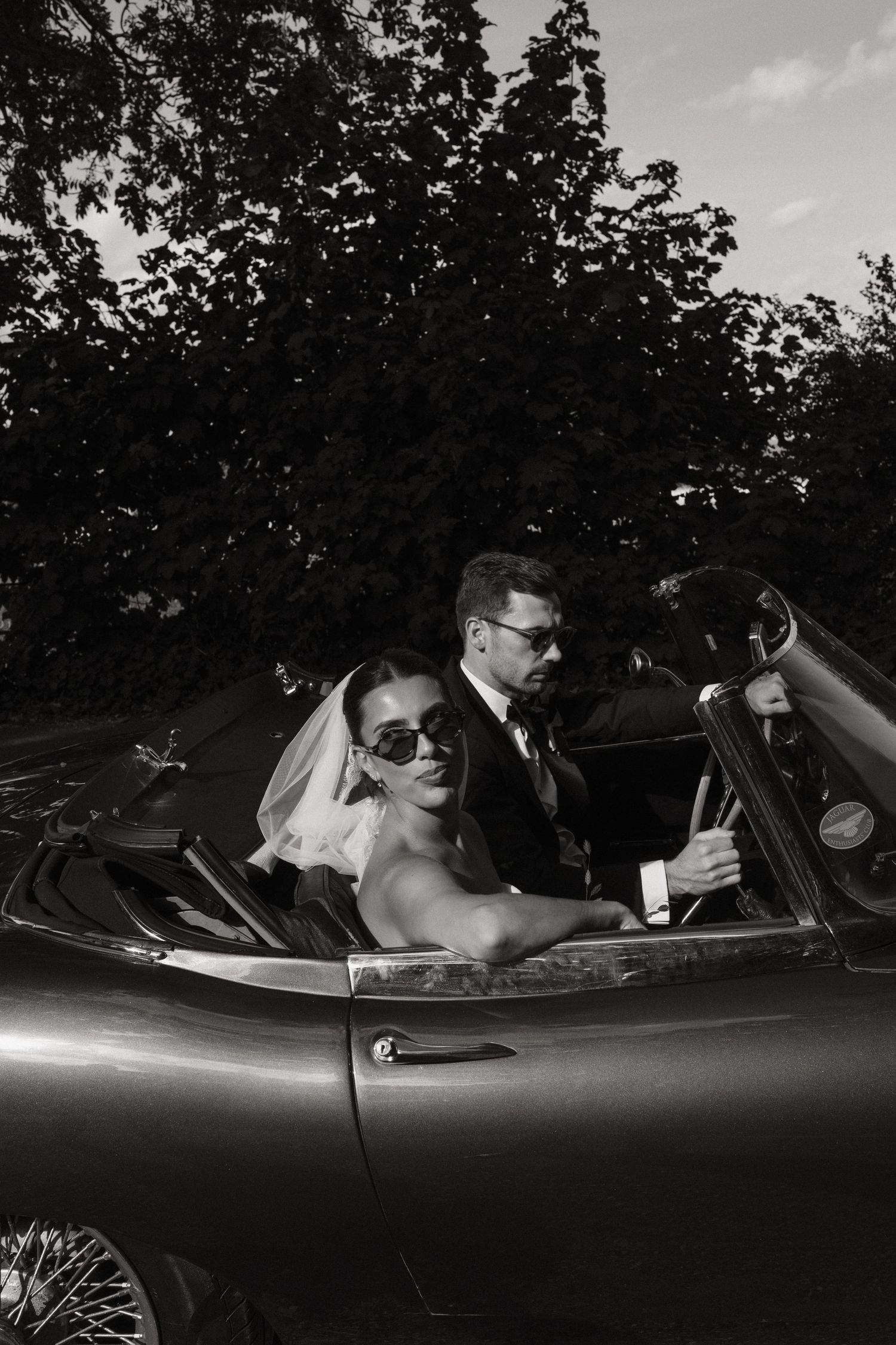 Black and white photo of a bride and groom in sunglasses riding in a classic convertible sports car on their wedding day.
