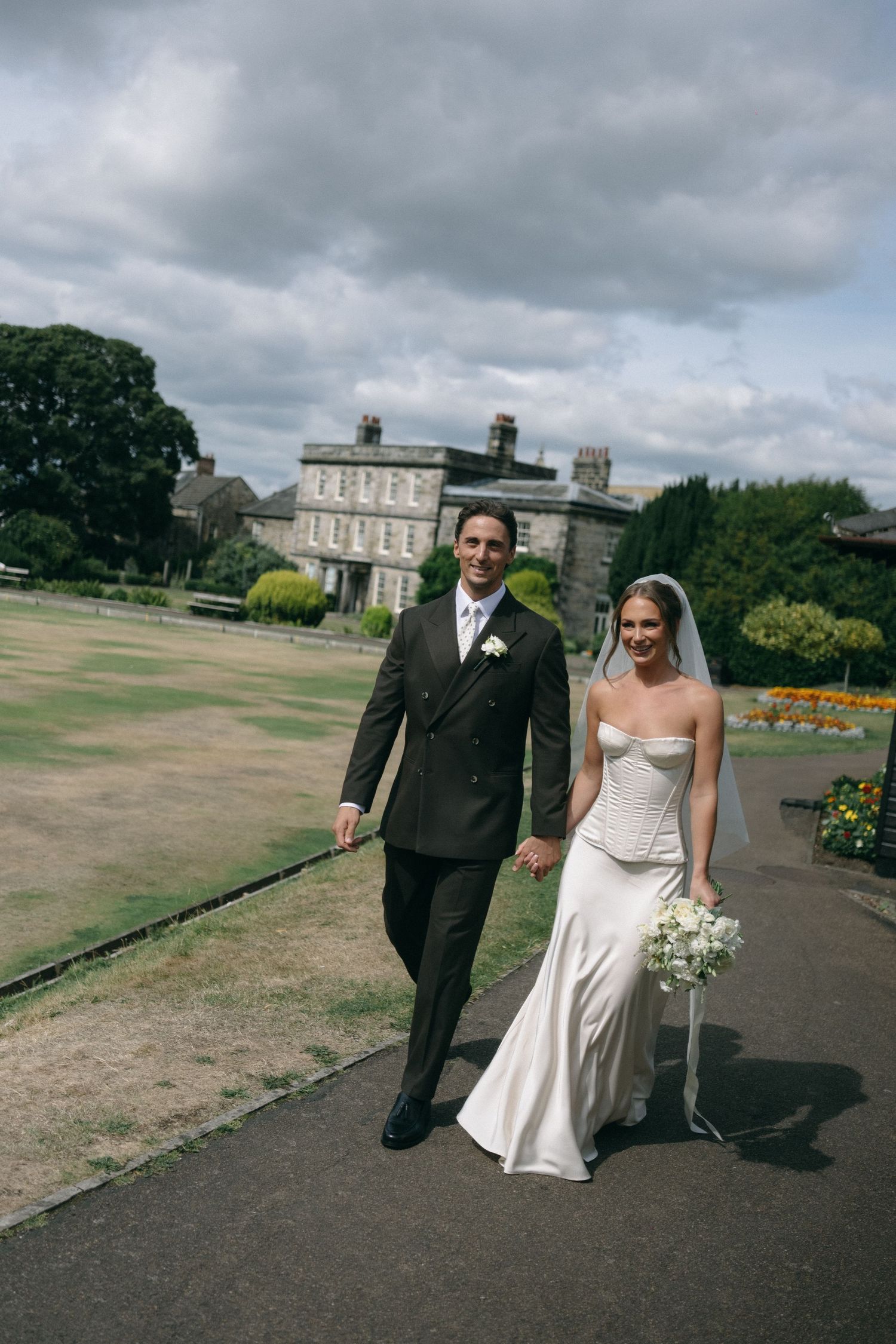 A bride and groom walk hand-in-hand on a garden path before a grand estate on their wedding day.