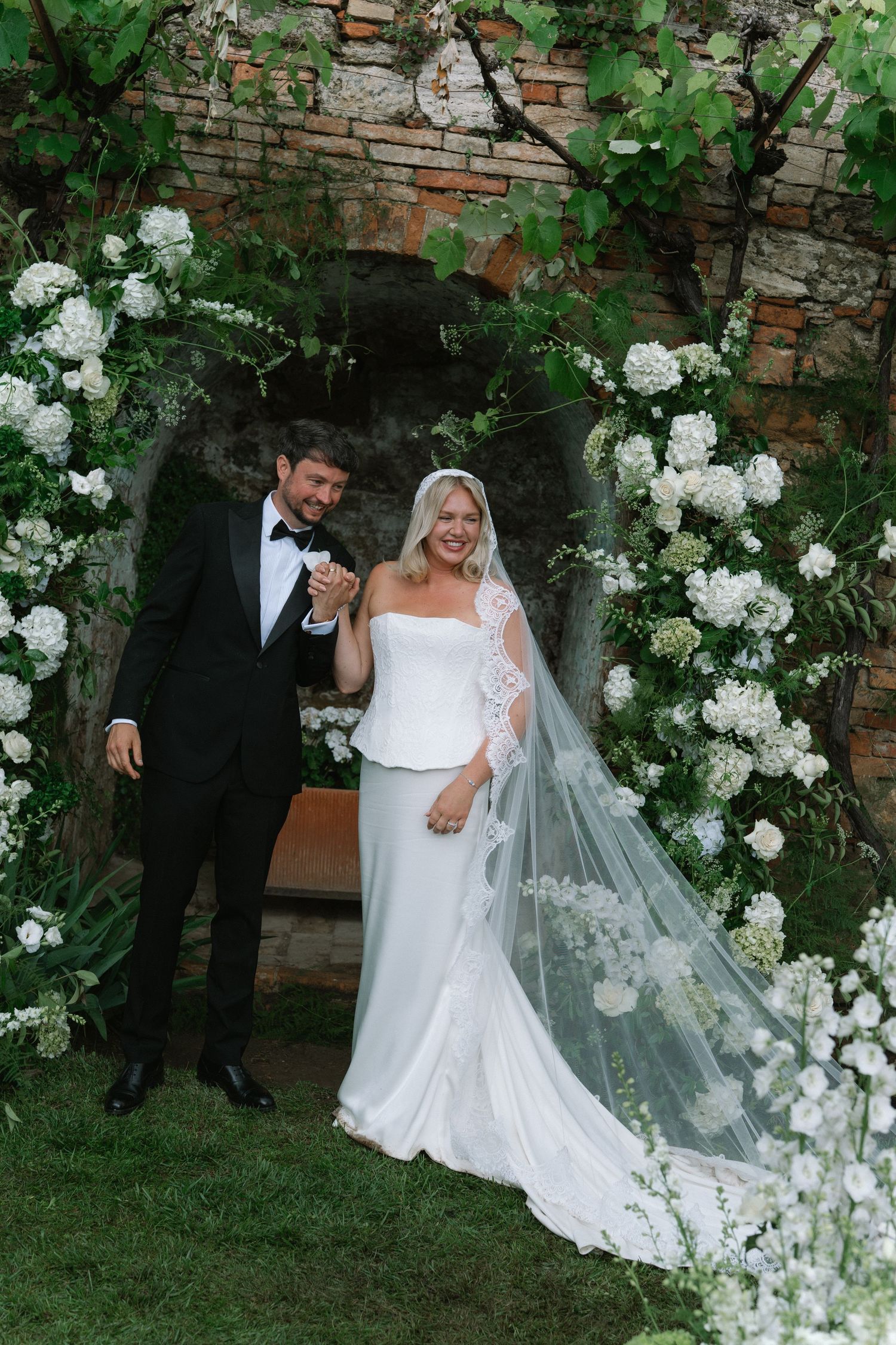 Smiling newlyweds pose together under a white floral arch at their outdoor garden wedding.