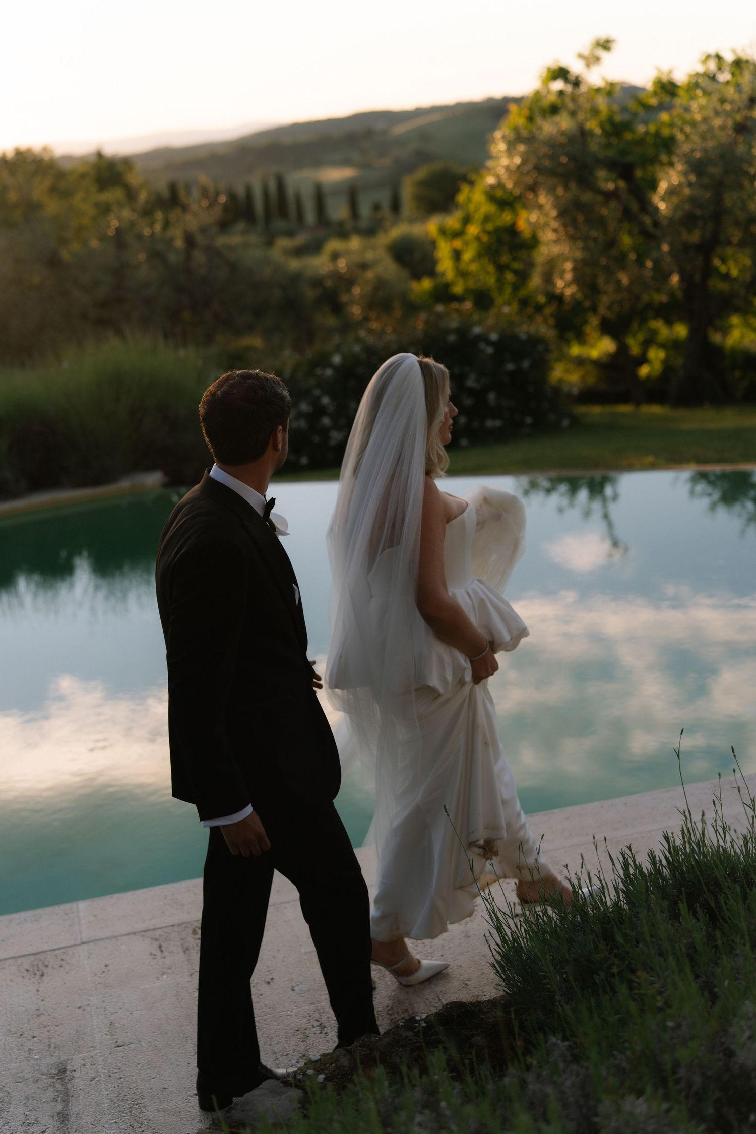 Bride and groom walk beside infinity pool reflecting sky at golden hour in Tuscany.
