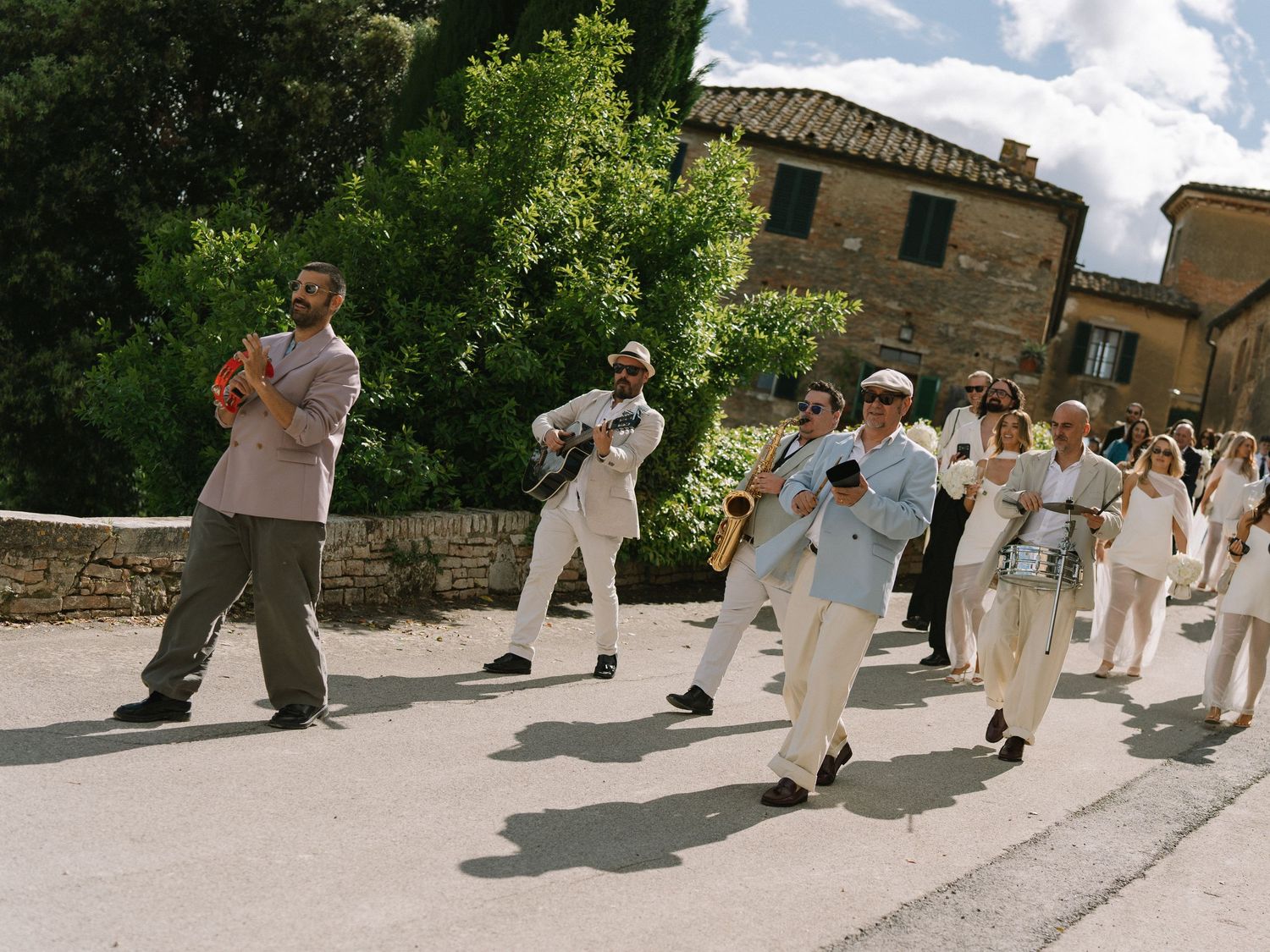 A lively brass band in white outfits performs outdoors on a sunny Tuscan road during a wedding procession.