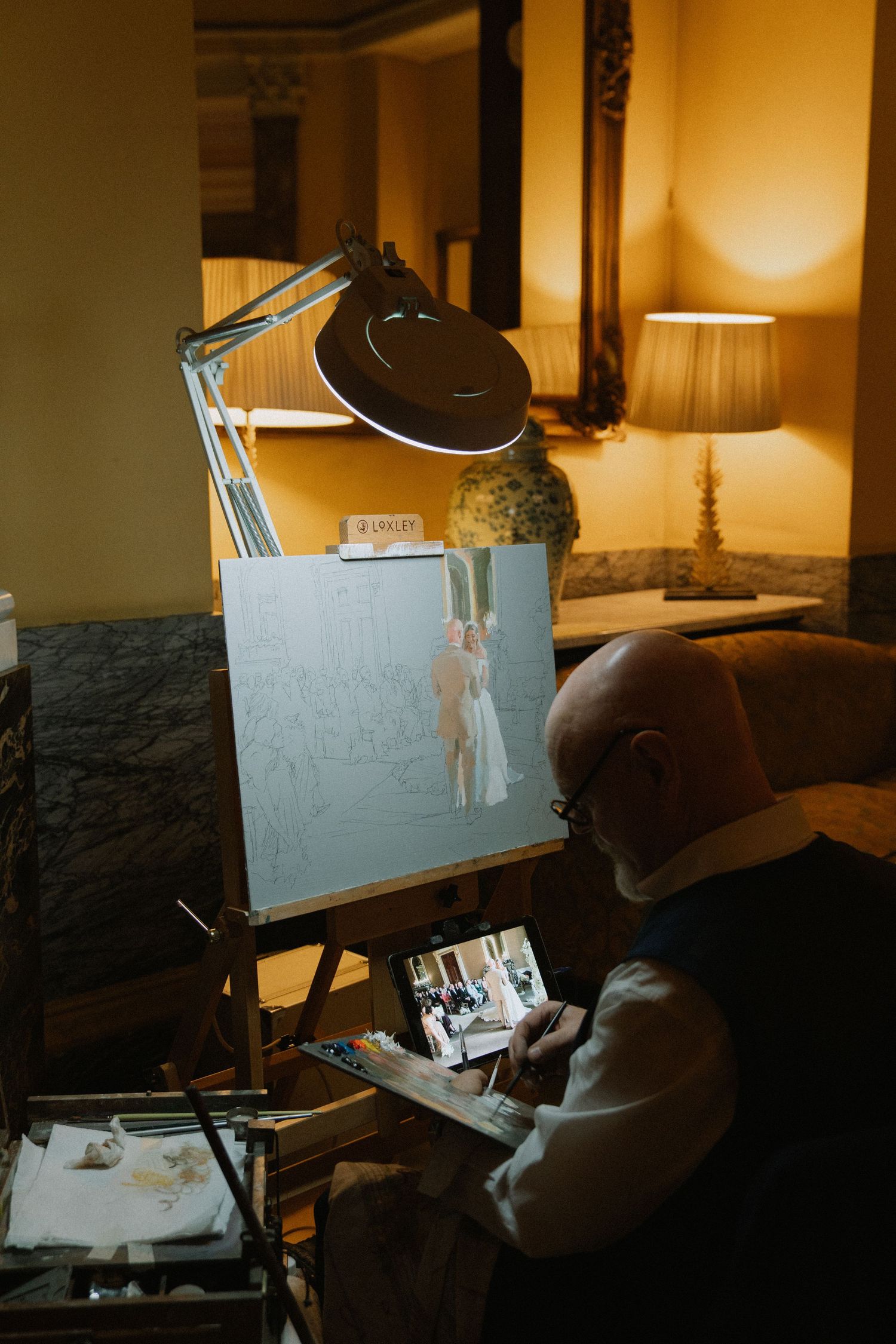 Artist painting a wedding portrait on an easel using a tablet reference, illuminated by a magnifying lamp in a warmly lit room.