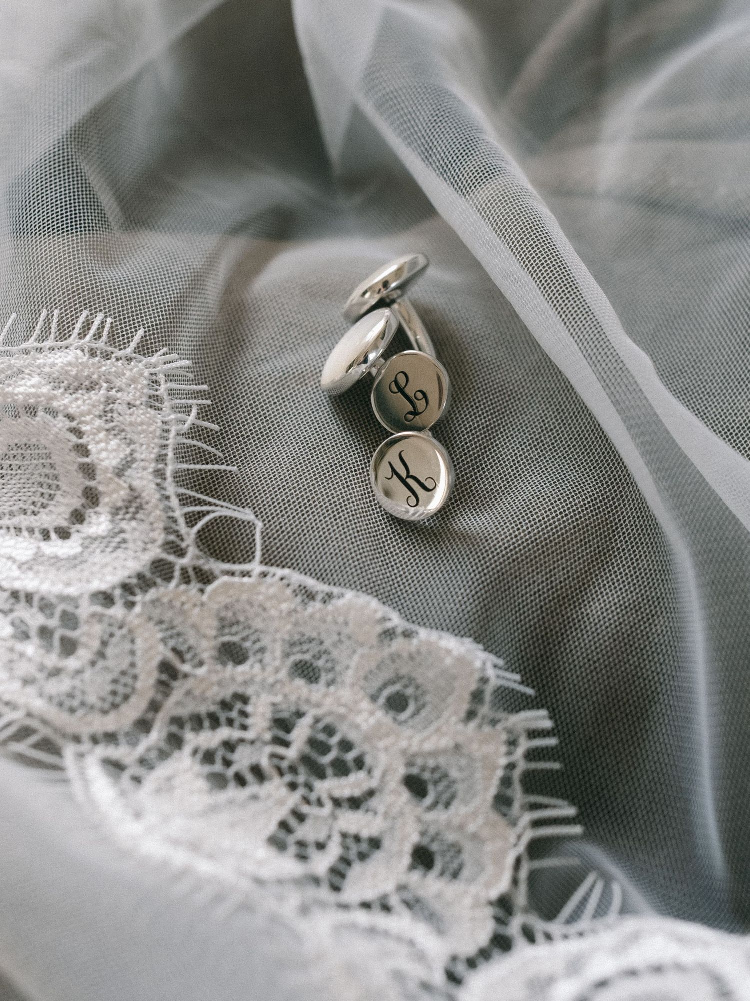 Silver monogrammed cufflinks resting on a delicate white lace wedding veil with soft gray tulle background.