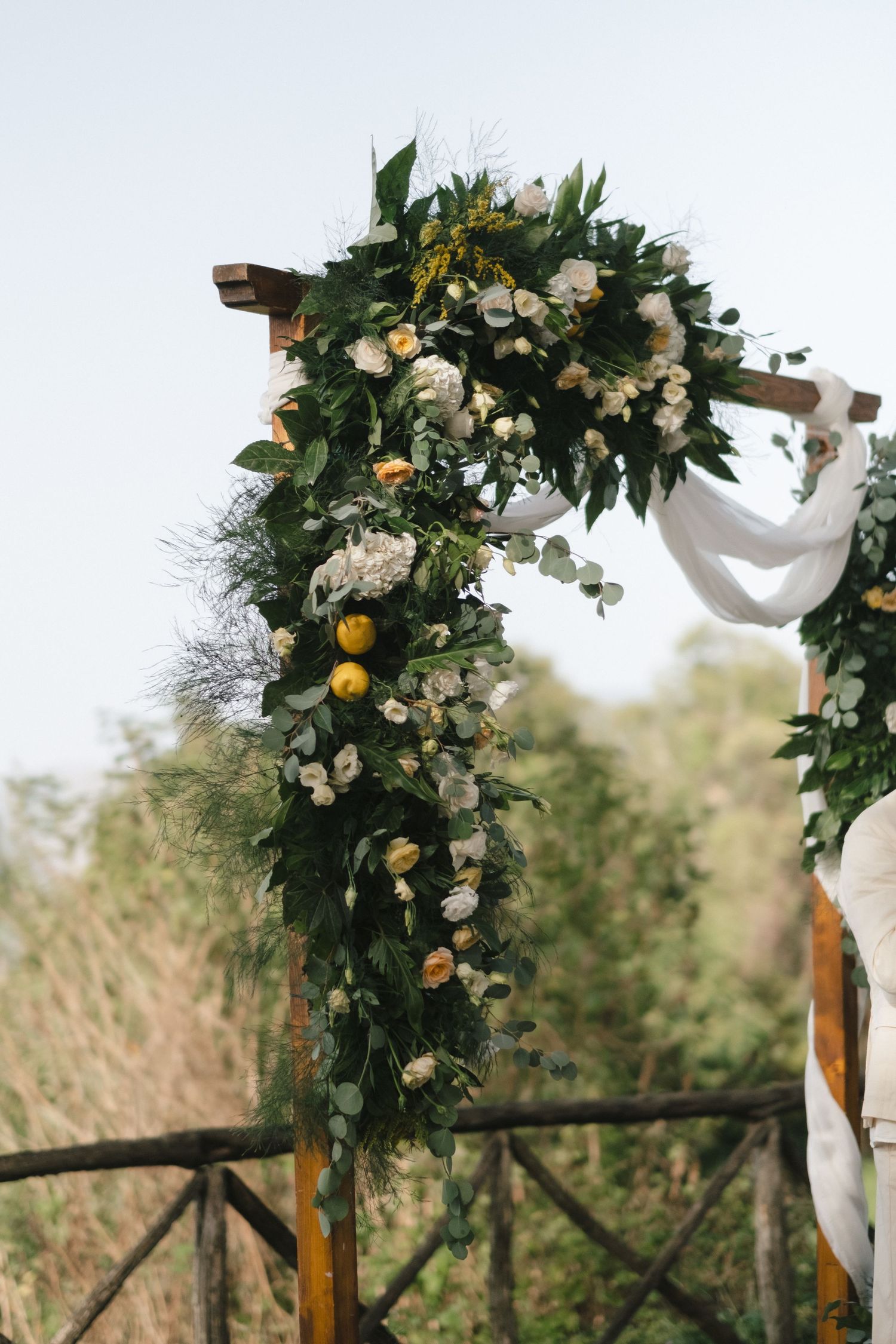 Rustic wooden wedding arch adorned with lush greenery, white roses, and yellow citrus fruit decorations.