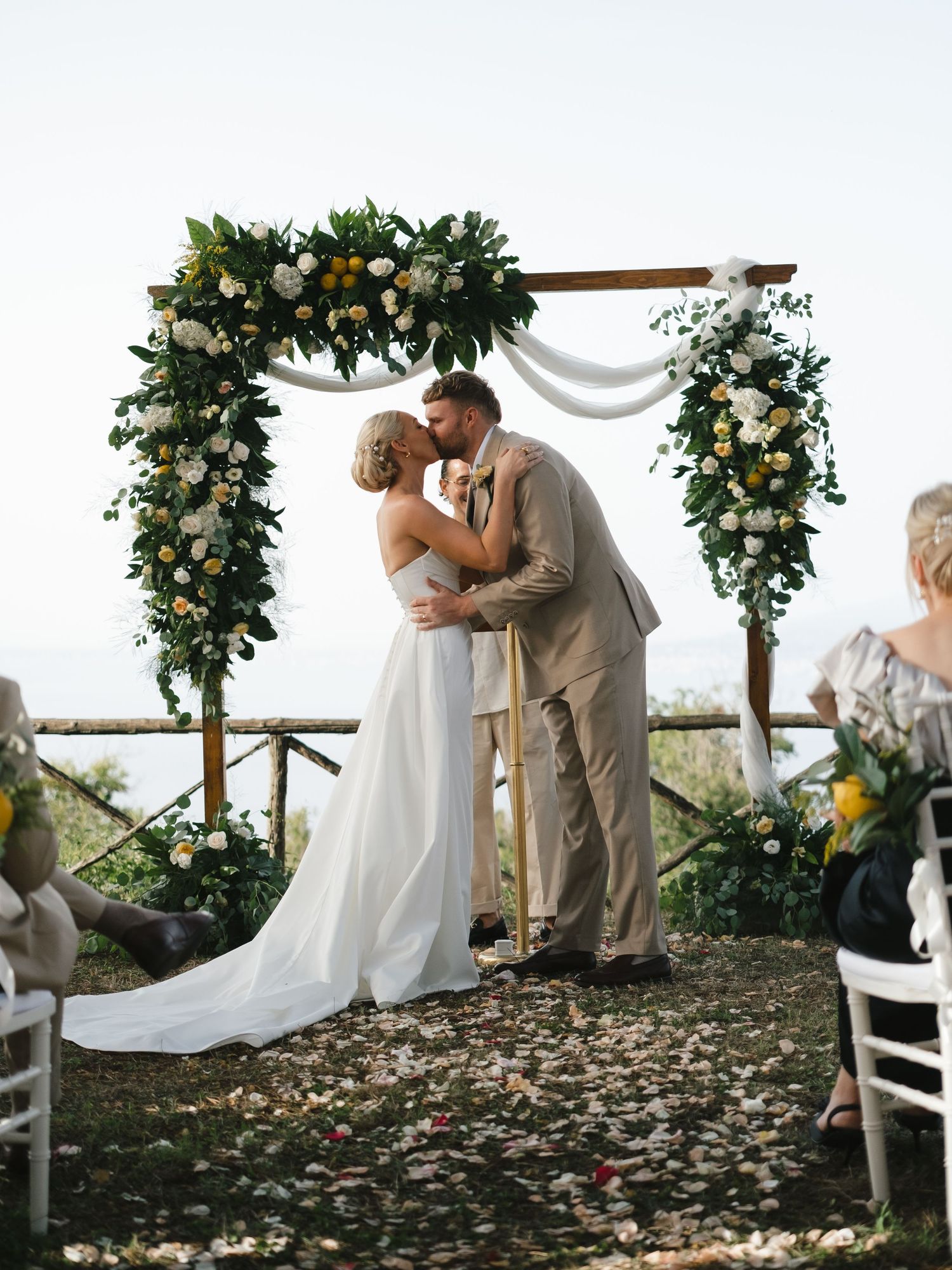 Bride and groom share first kiss under a lush greenery and white floral wedding arch with ocean views.