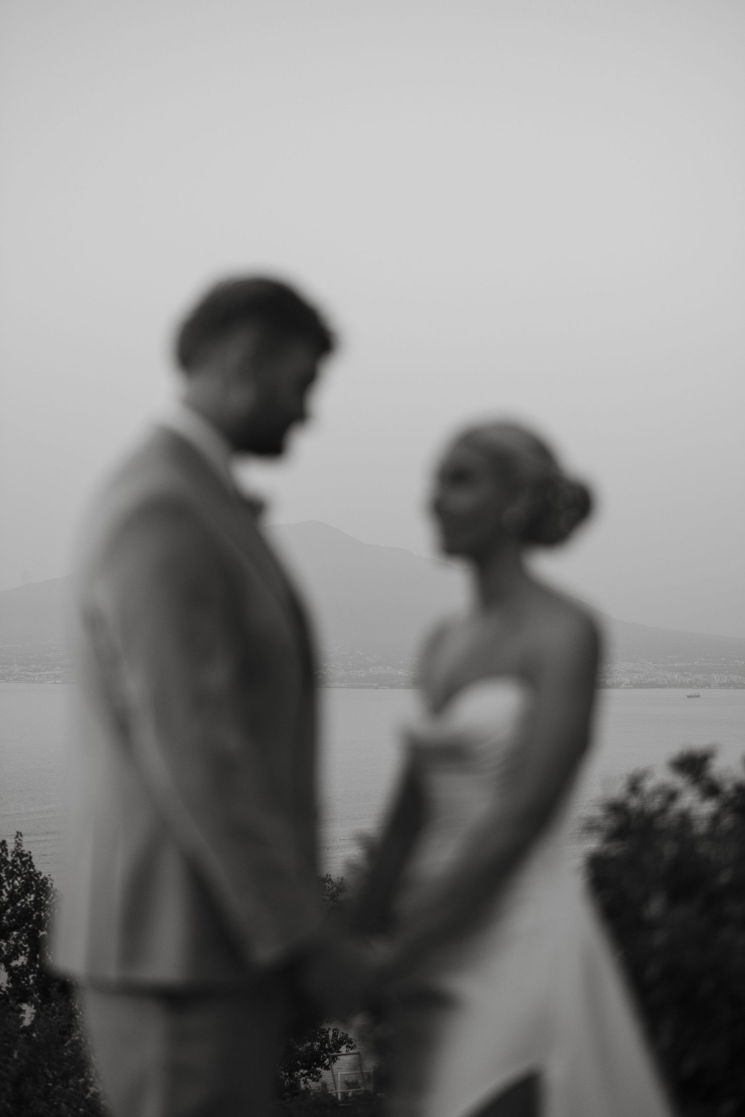 Blurred black and white portrait of bride and groom holding hands with mountain and lake backdrop.