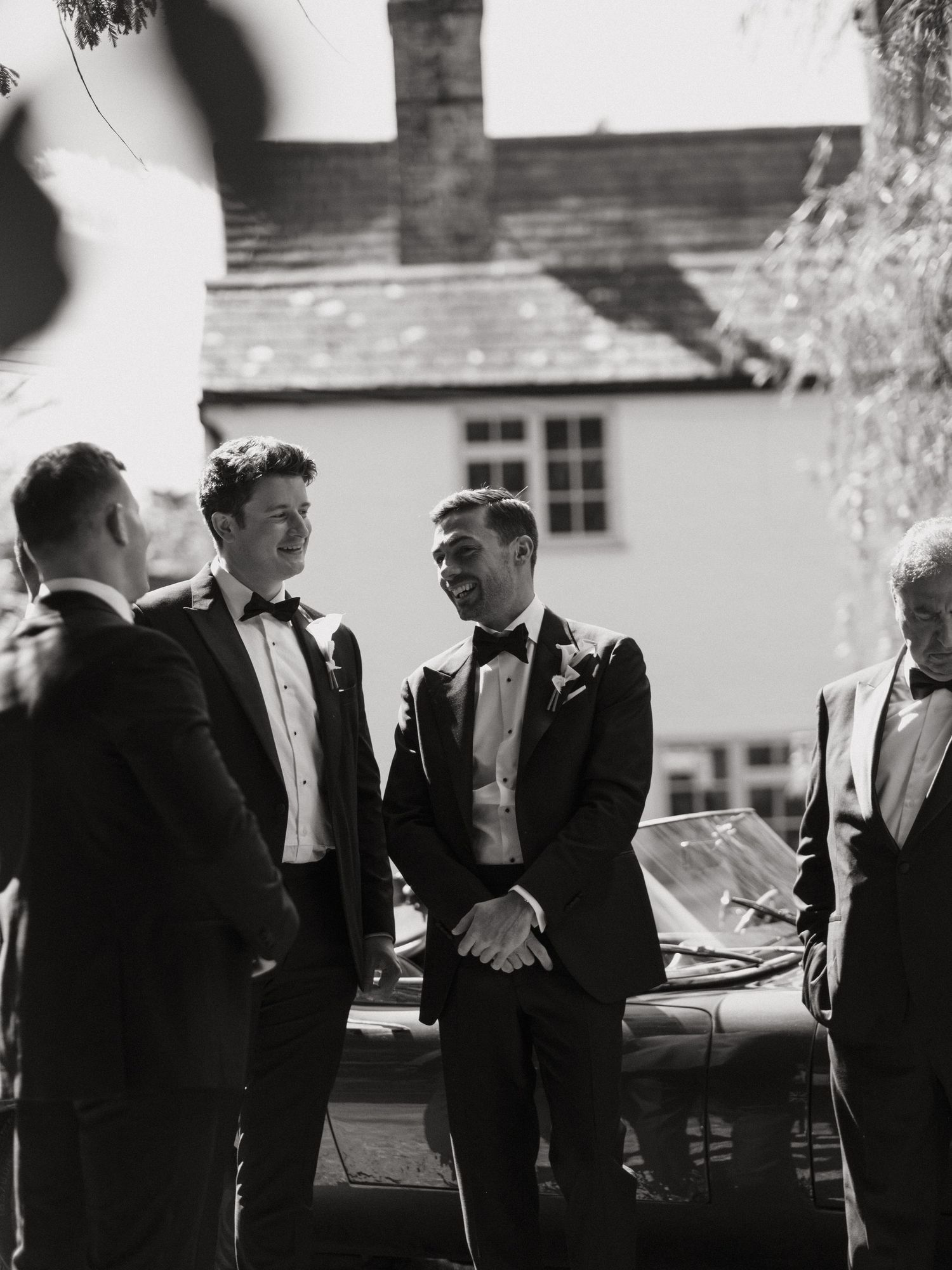 Groomsmen in black tuxedos laughing together outdoors at a wedding venue in a black and white photo.