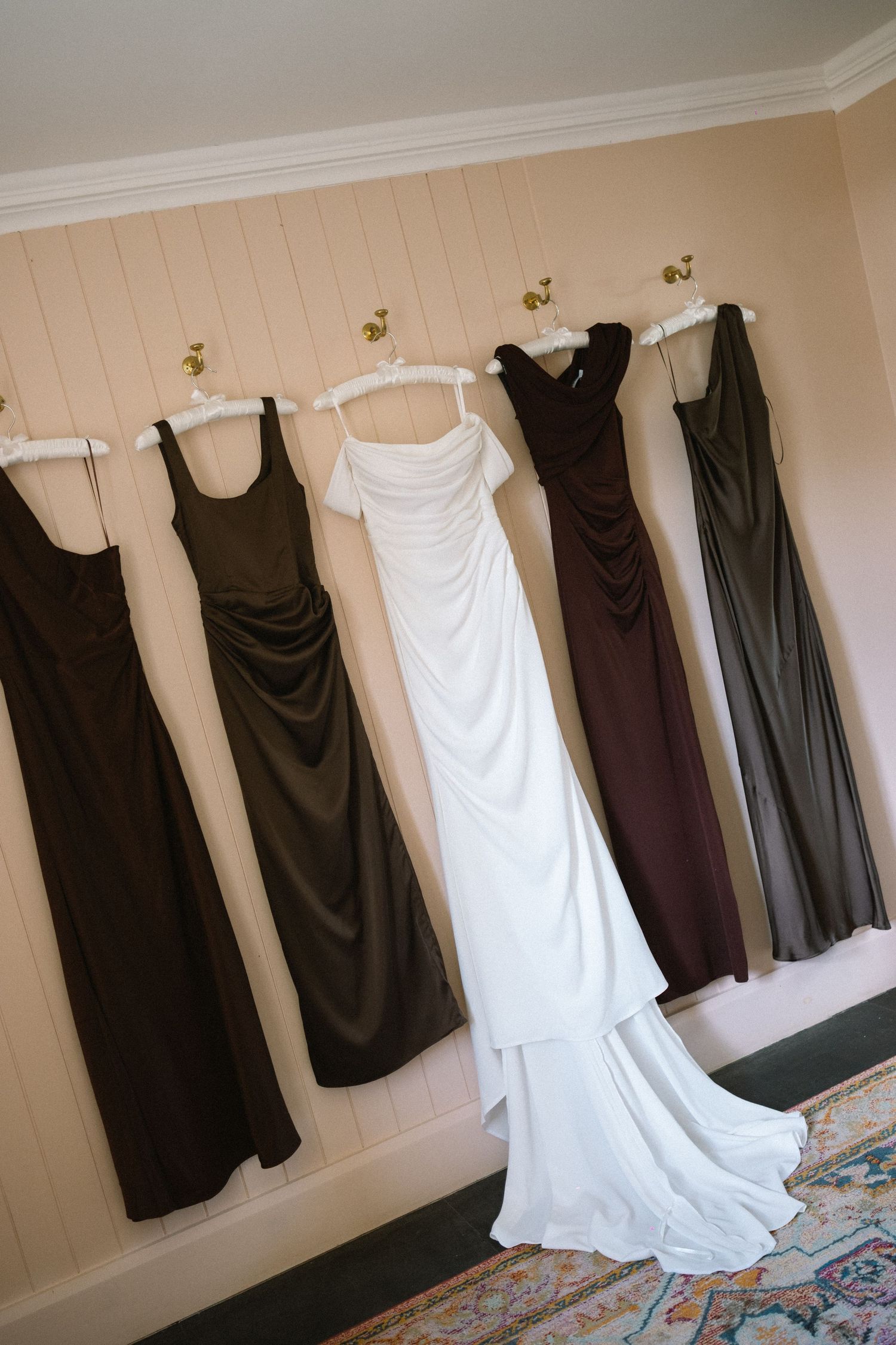 Five formal gowns hanging on wall hooks, including a white bridal gown flanked by dark bridesmaid dresses.