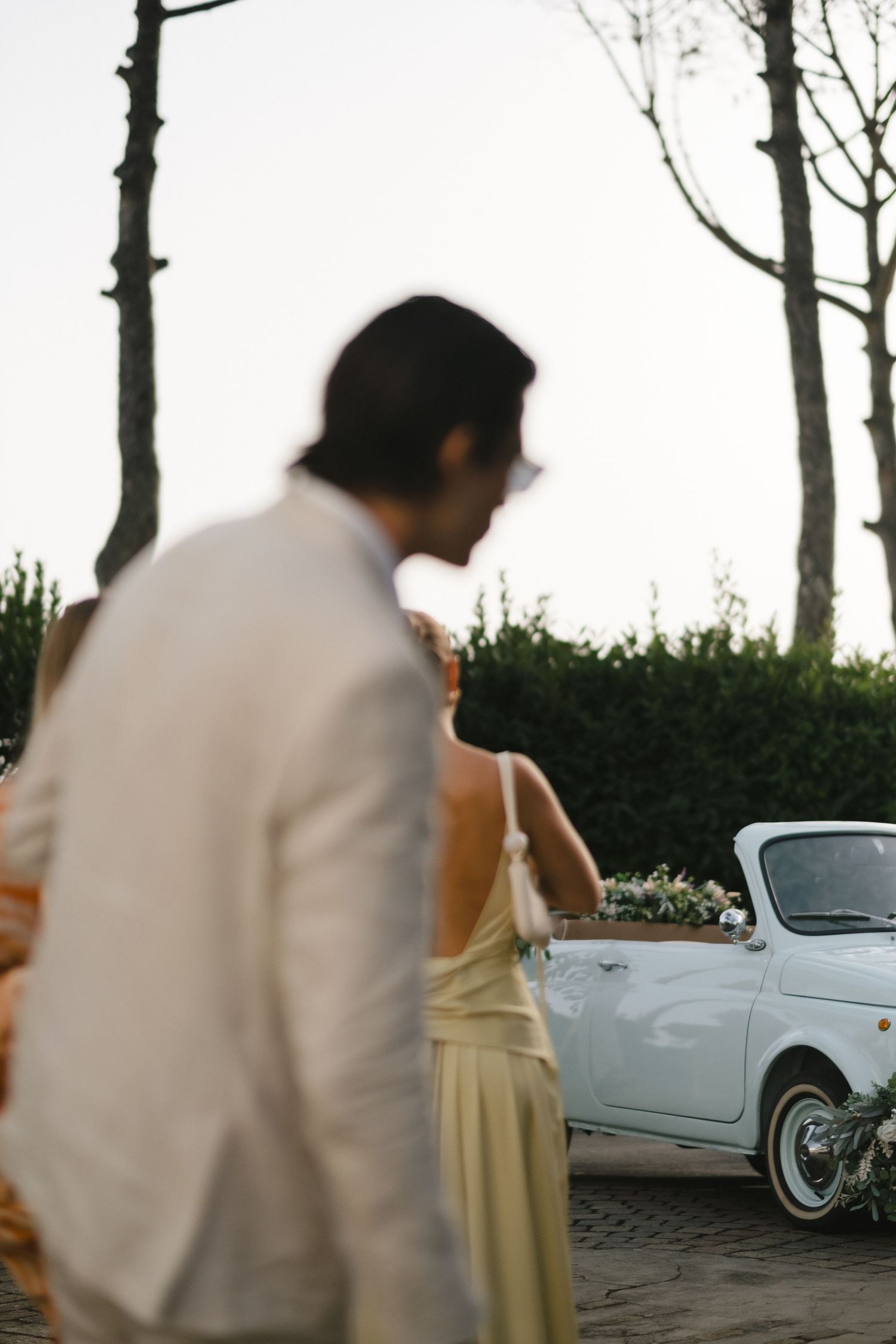 Blurred couple in foreground with vintage white Fiat 500 decorated with flowers at wedding reception.