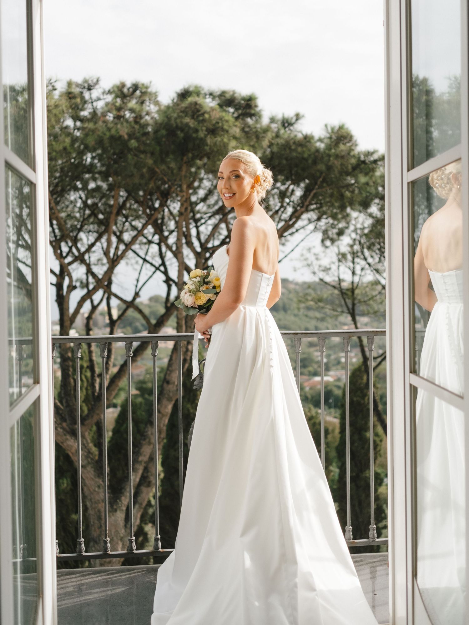 Elegant bride in strapless white gown holding bouquet on balcony with lush green trees in background.