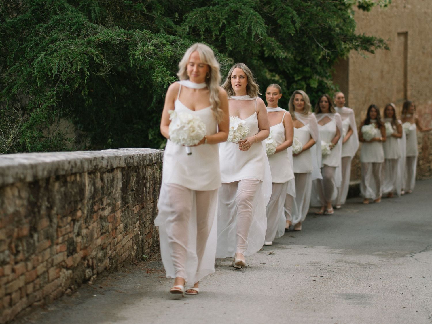 Long line of bridesmaids in matching white outfits carrying bouquets walking along a stone wall at a wedding.