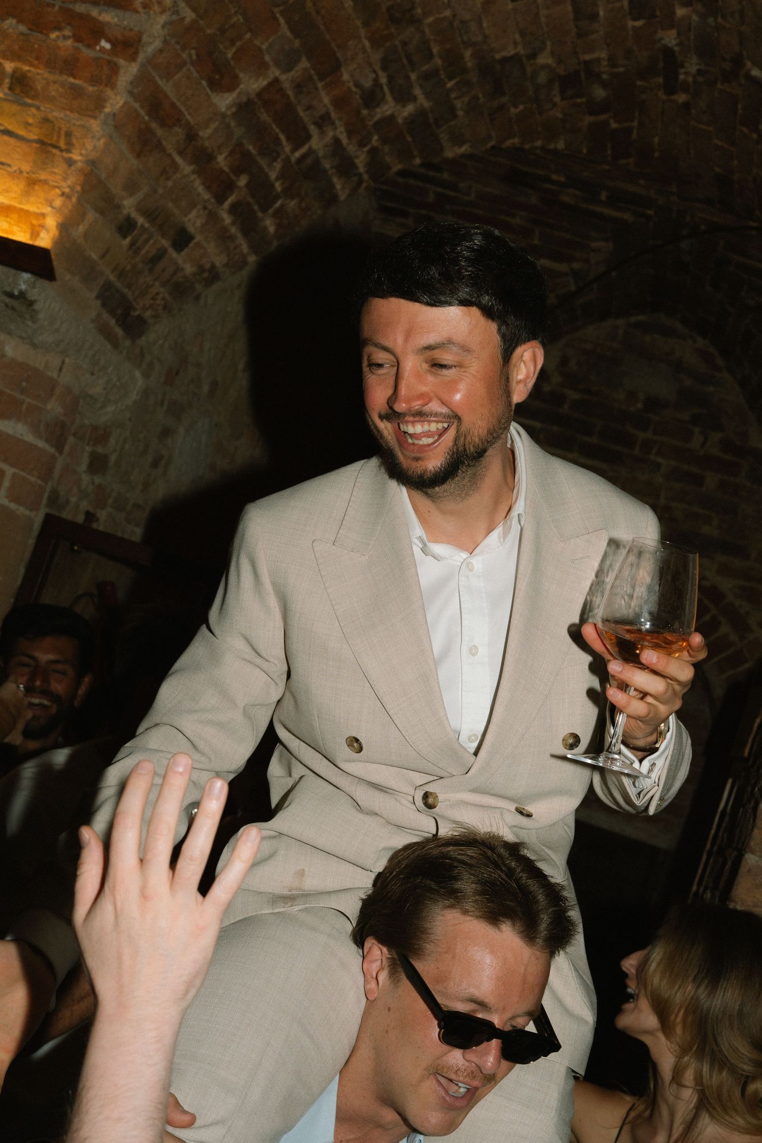 A laughing man in beige suit holds a champagne glass while being carried on someone's shoulders at a wedding party.