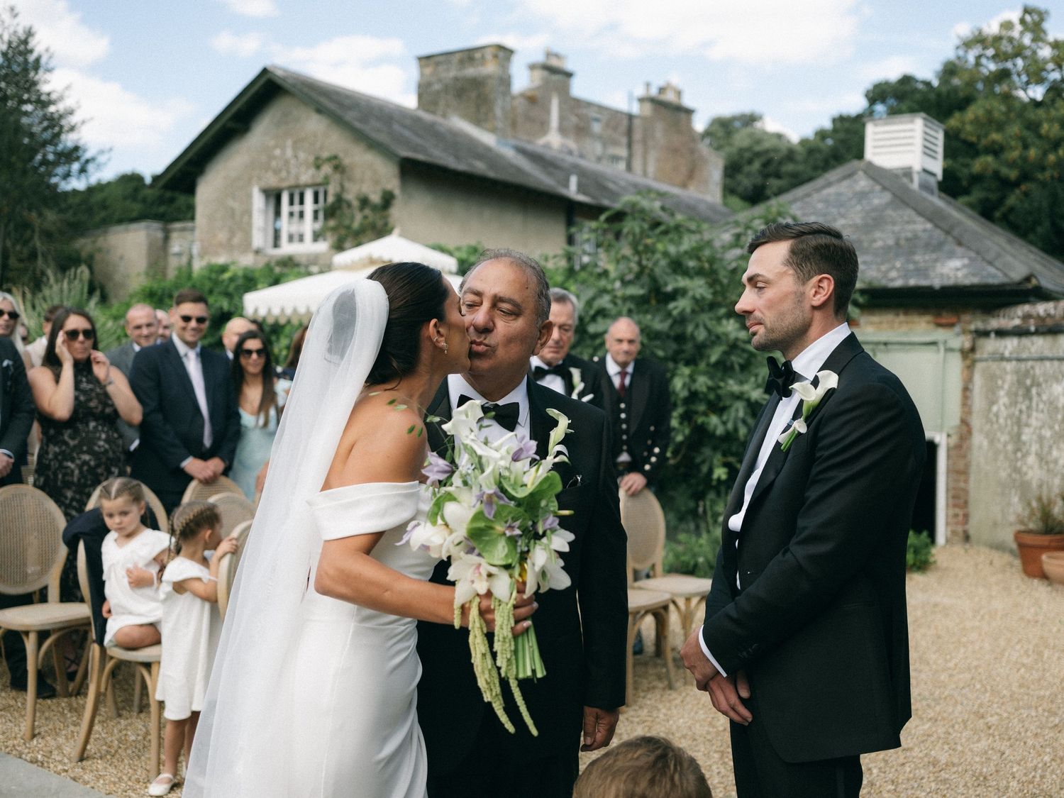 Bride and groom share a tender moment at outdoor wedding ceremony with guests seated in garden of historic stone manor.