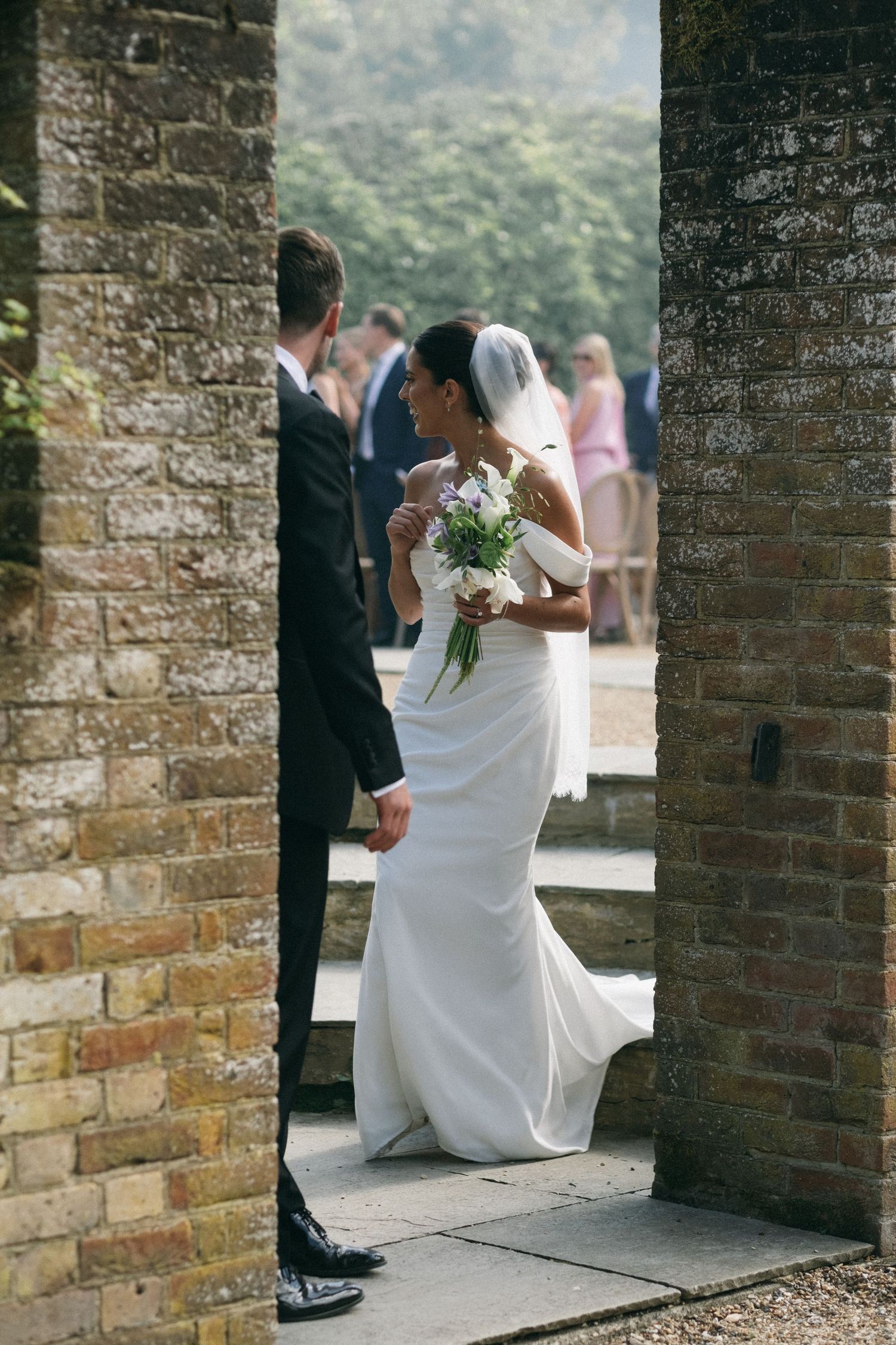 Bride in white gown holding bouquet stands with groom in black suit between stone pillars at outdoor wedding venue.