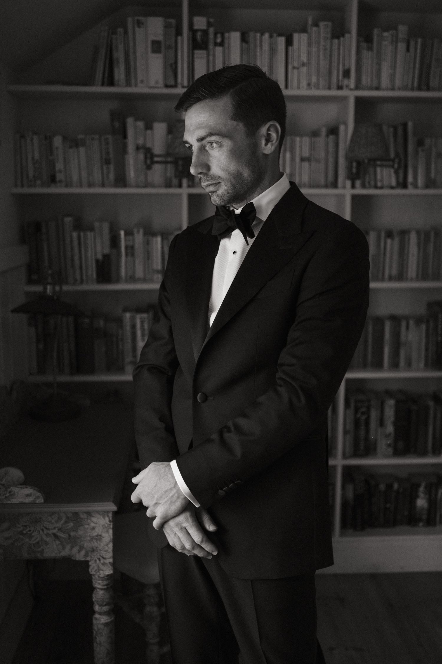 Elegant man in black tuxedo and bow tie poses in front of bookshelf in moody black and white portrait.