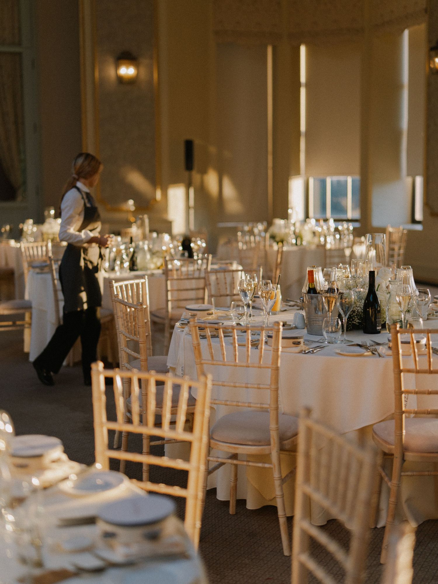 Elegant banquet hall with gold chiavari chairs, white linen tables, and a server preparing for a formal wedding reception.