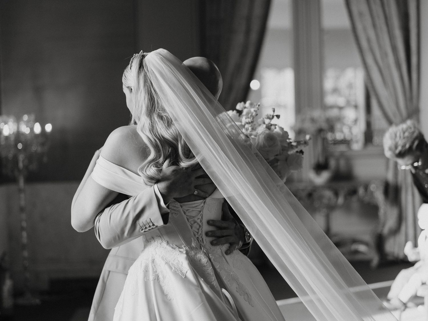 Black and white photo of a bride in a veil embracing someone at her wedding reception indoors.